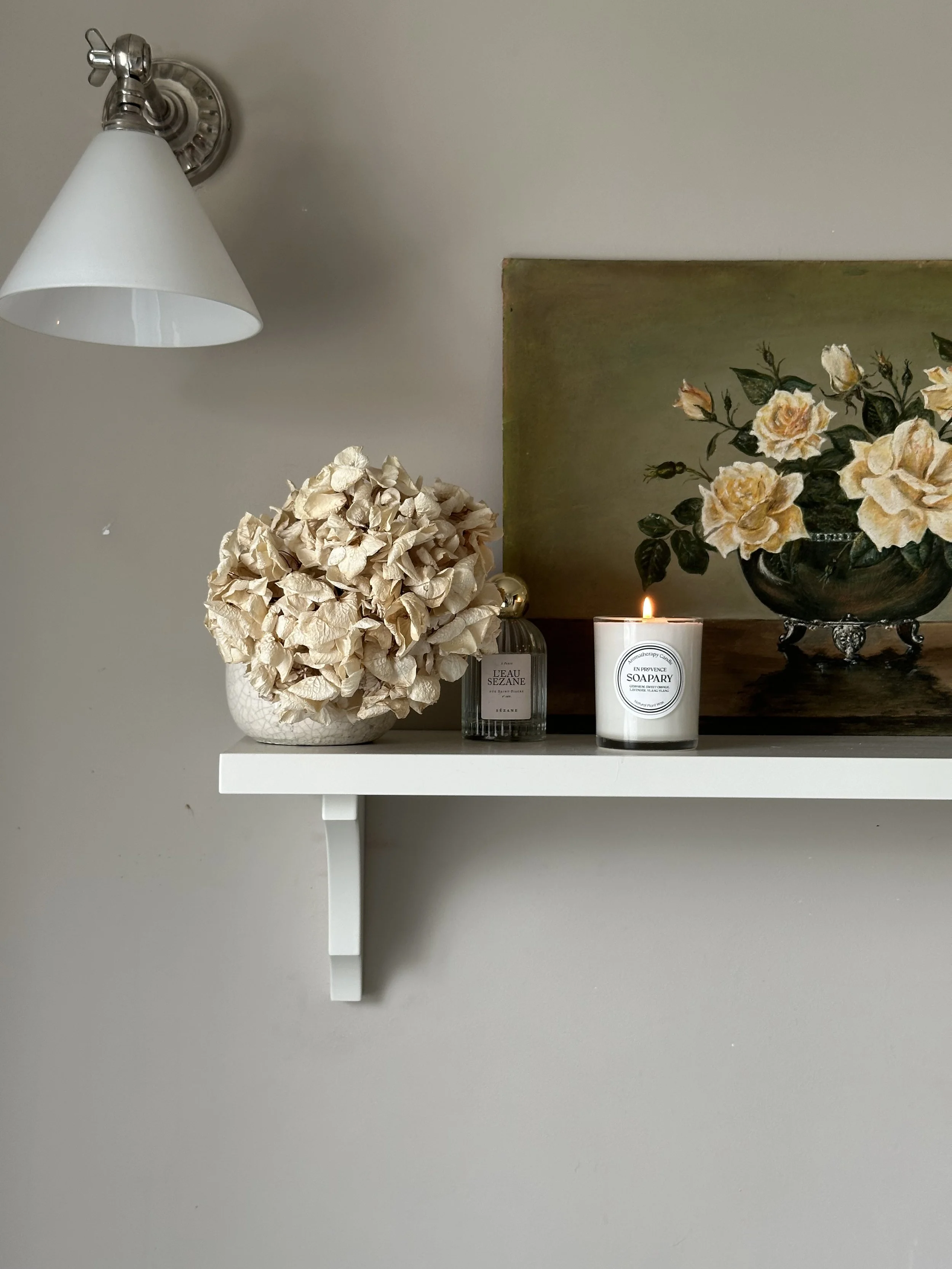 A small white shelf mounted to a light gray wall, holding a white hydrangea floral arrangement, a lit white candle, a bottle of perfume, and a decorative tray with yellow roses painting in the background.