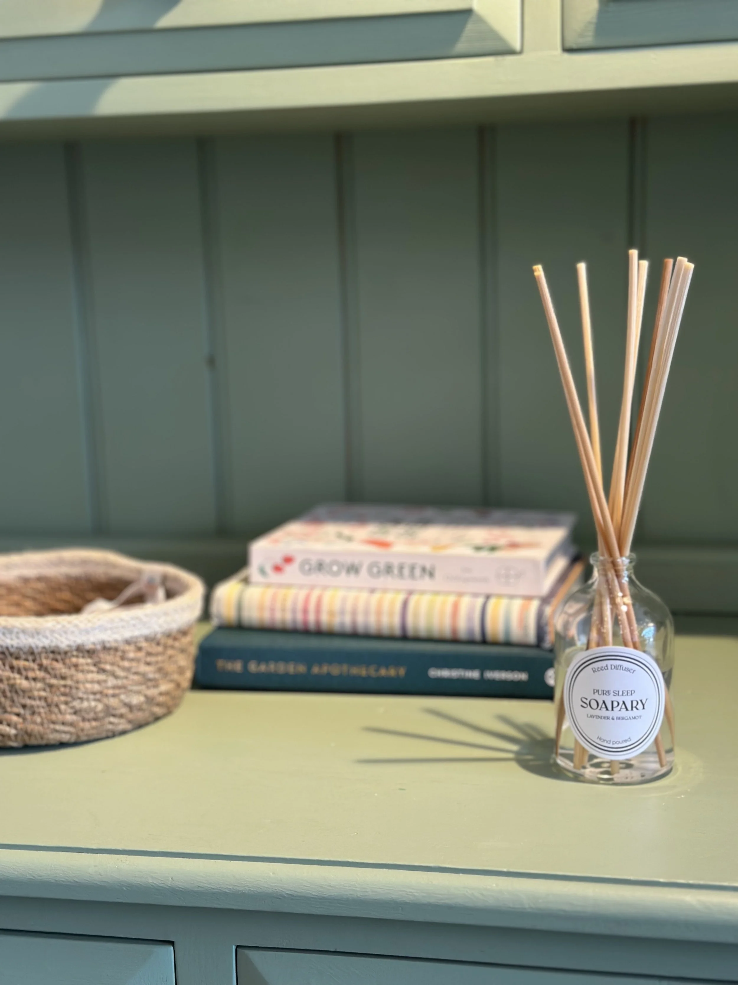 A green desk with a glass jar of reed diffusers, a woven basket, and books about gardening and herbs.