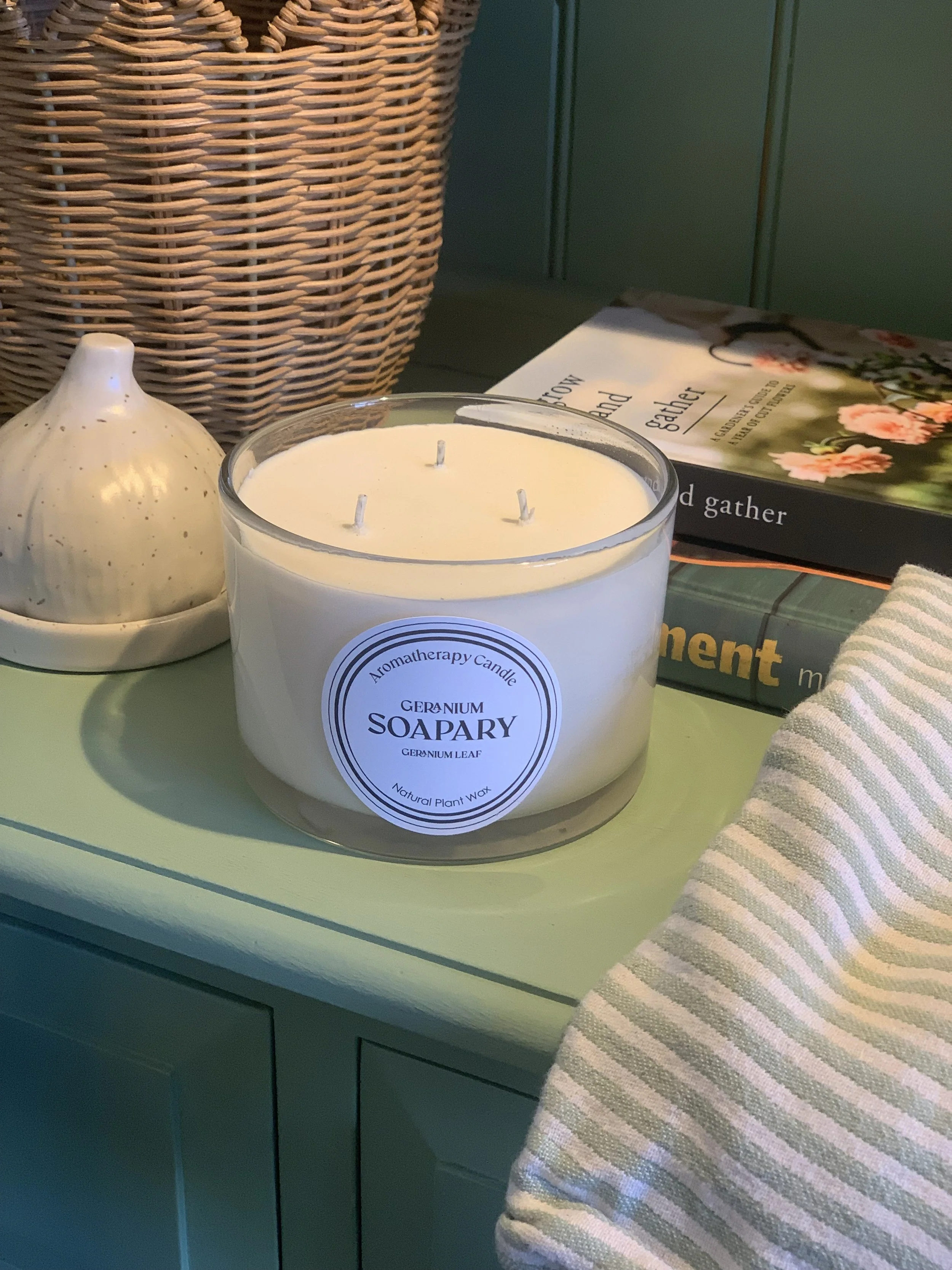 A white aromatherapy candle labeled 'Geranium Soapary' with three wicks, placed on a light green table. Next to the candle is a small white ceramic object and a book titled 'Gather' with floral cover art. A striped cloth is partially visible in the foreground, and a wicker basket is in the background.