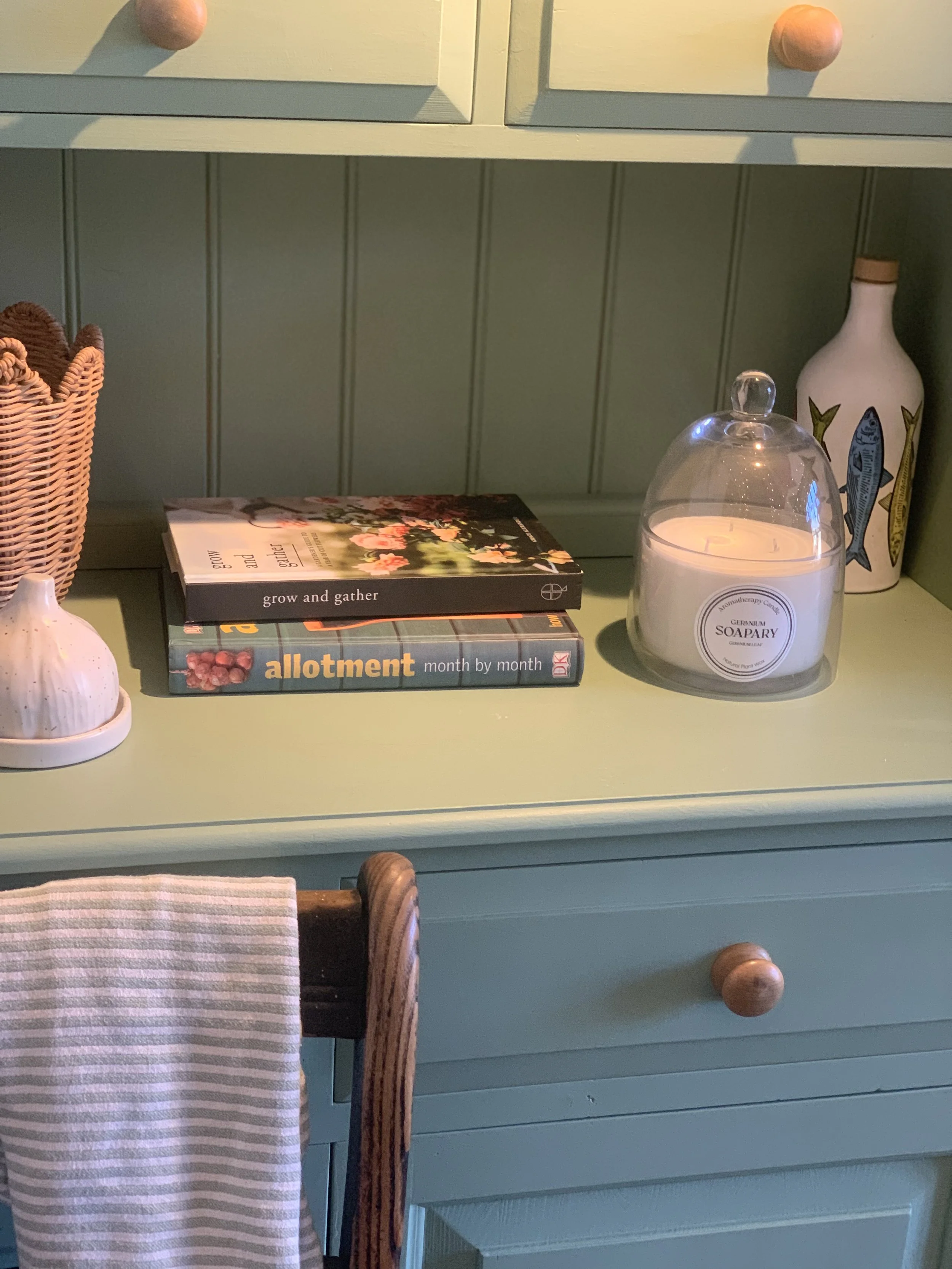 A green wooden sideboard with stacked magazines, a lit white candle in a glass cover, a ceramic vase with fish illustrations, a wicker basket filled with small objects, and a small white ceramic knickknack.