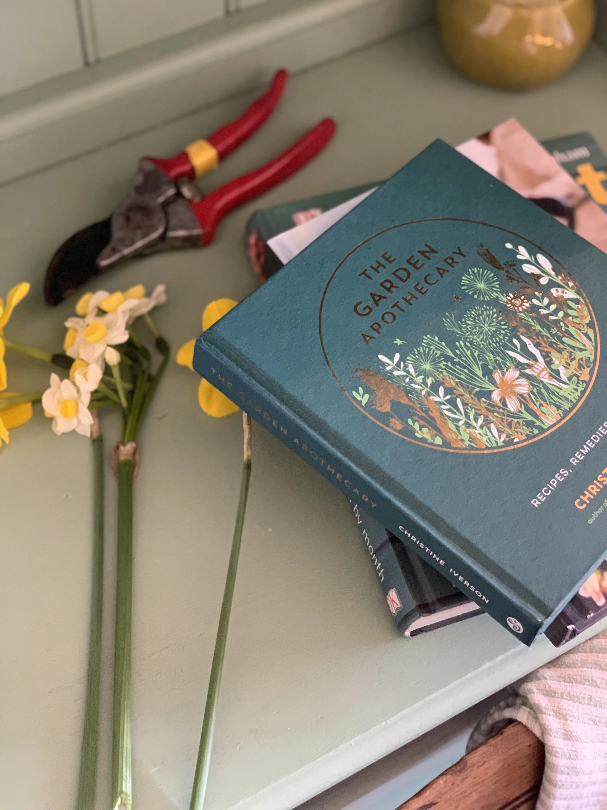 Close-up image of a stack of books titled 'The Garden Apothecary' with some yellow and white flowers beside them, a pair of pruning shears, and part of a pink striped blanket or towel on a light-green surface.