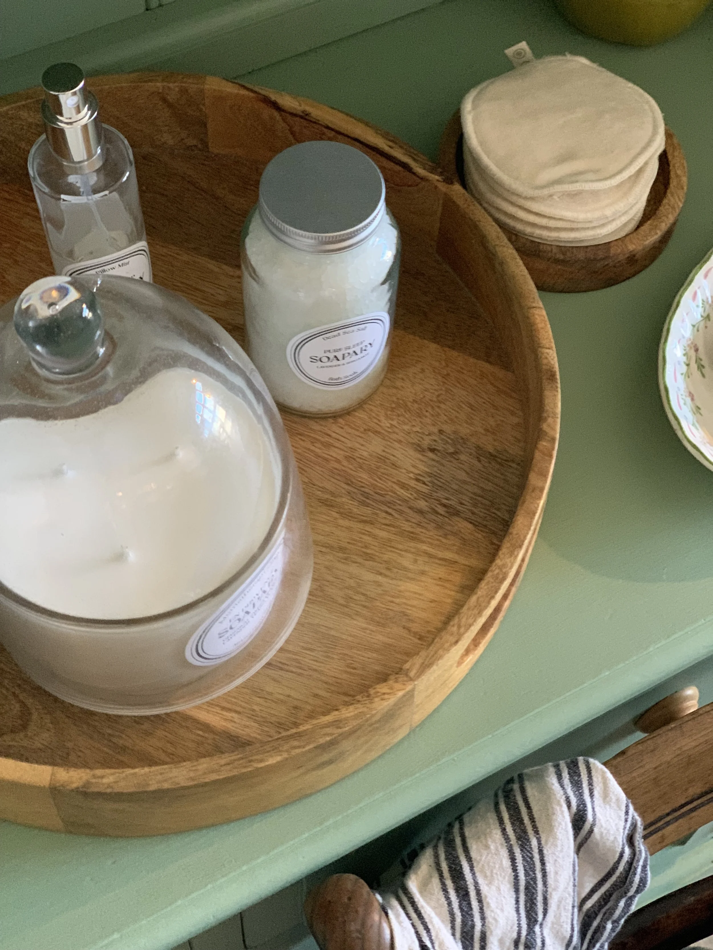 A wooden tray with a large white candle, a bottle of hand sanitizer, and a jar of soap flakes on a green surface. There are also white reusable facial rounds and a decorative plate with a floral pattern nearby.