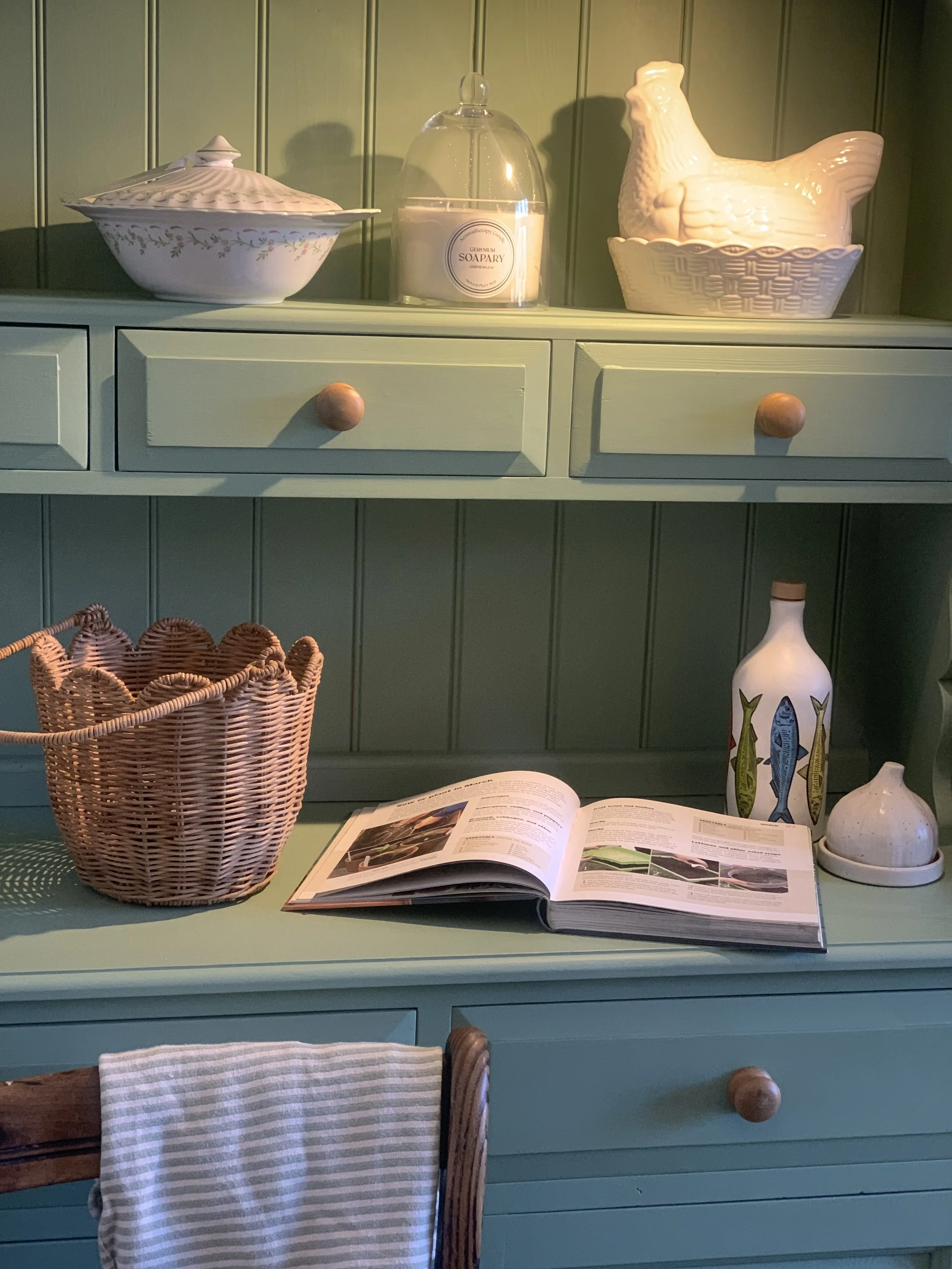 A green wooden hutch with decorative plates, soap dish, and ceramic chicken figurine on top. The lower surface has a wicker basket, an open magazine, a ceramic bottle with fish design, and a small ceramic garlic bulb. A striped towel hangs from a wooden chair in front.