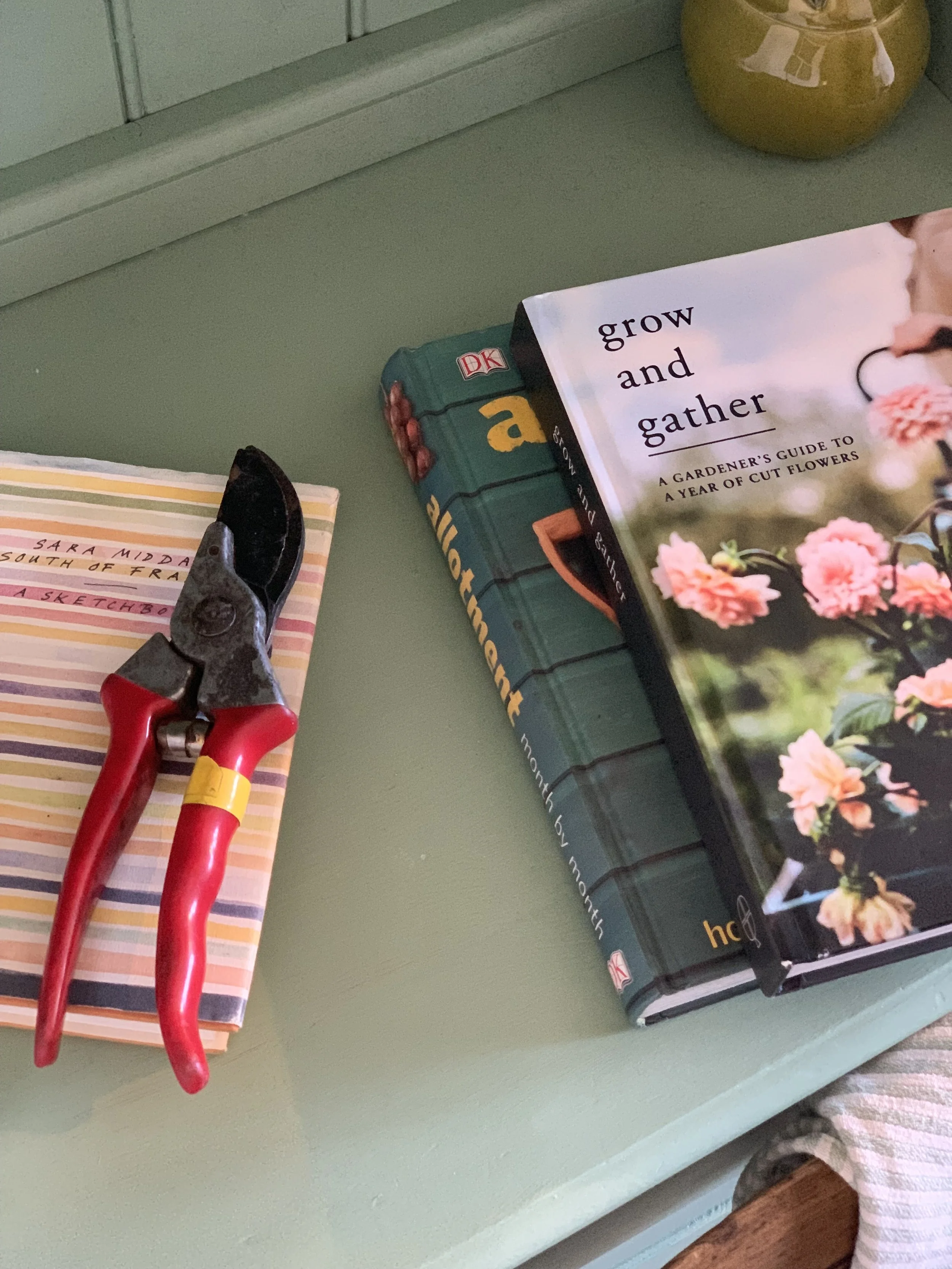 A green bookshelf with a pair of red-handled pruning shears, a book titled 'grow and gather,' a striped notepad, and a yellow vase.