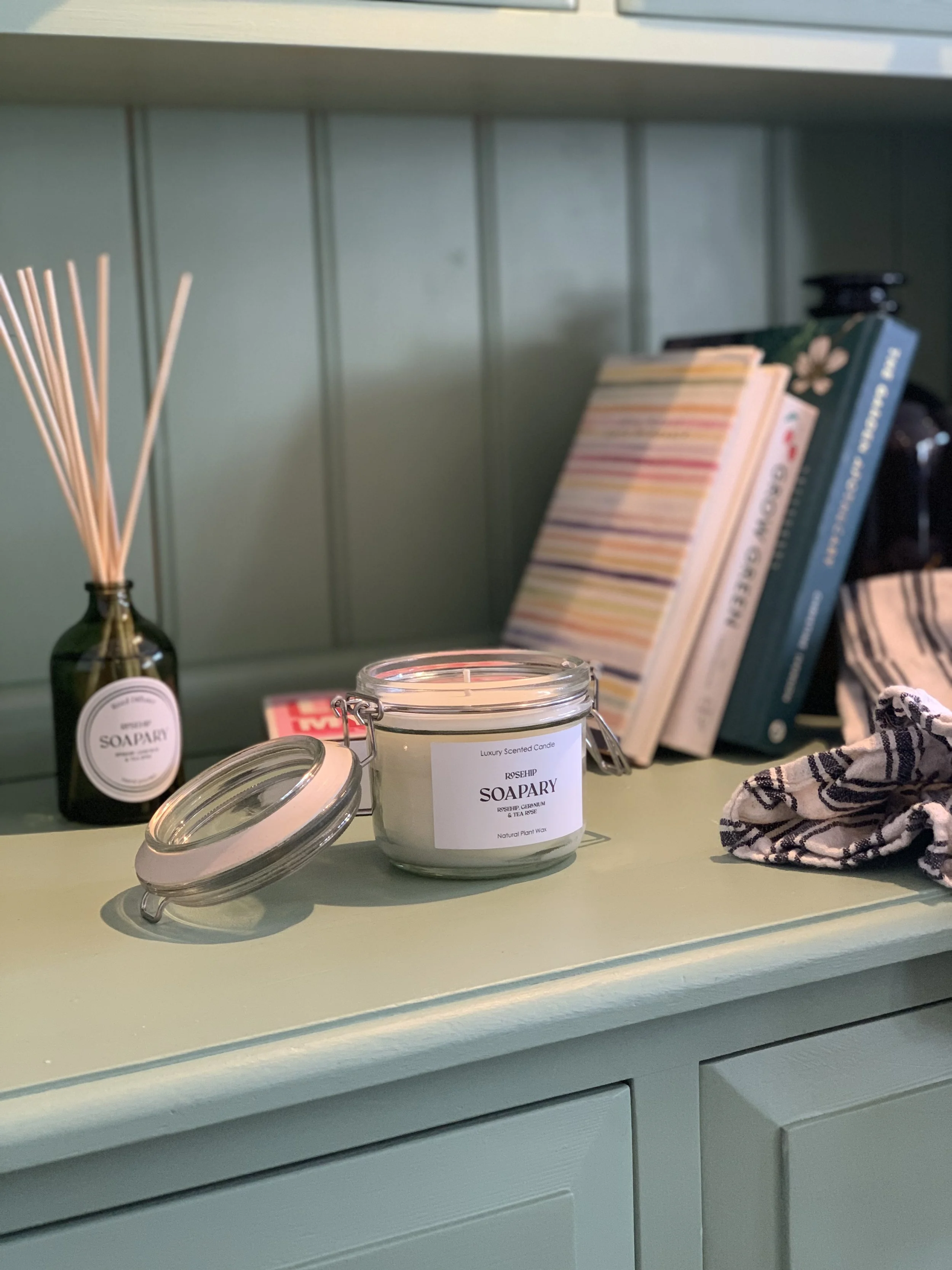 A collection of candles and home decor on a green shelf, including a jar candle labeled 'luxury scented candle,' a bottle with reed diffusers, a few books, a patterned cloth, and a dark object in the background.
