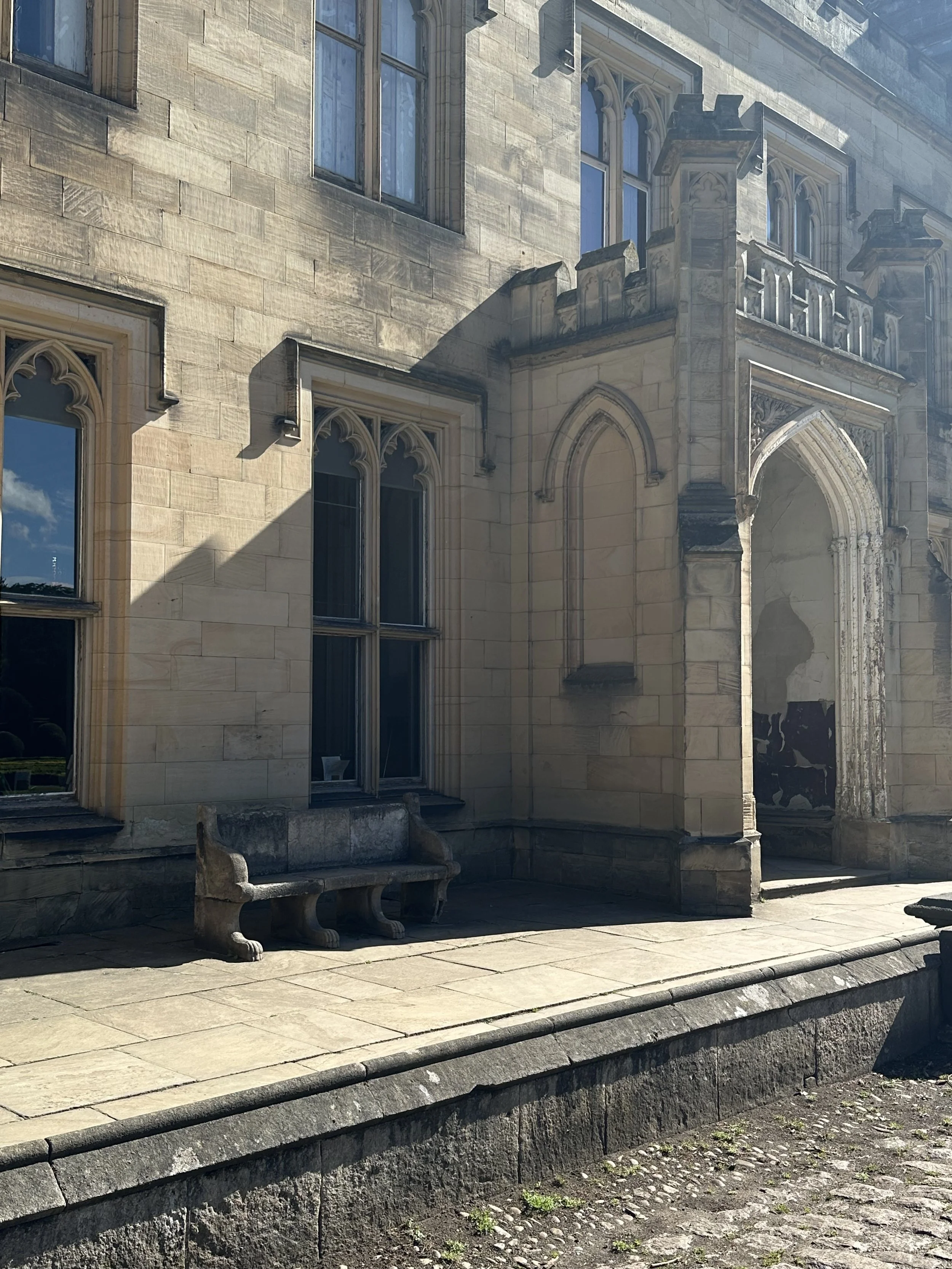 Ancient stone building with tall, pointed arched windows and decorative stonework. There are stone benches in front and a paved path along the building. Shadows cast on the building indicate a sunny day.