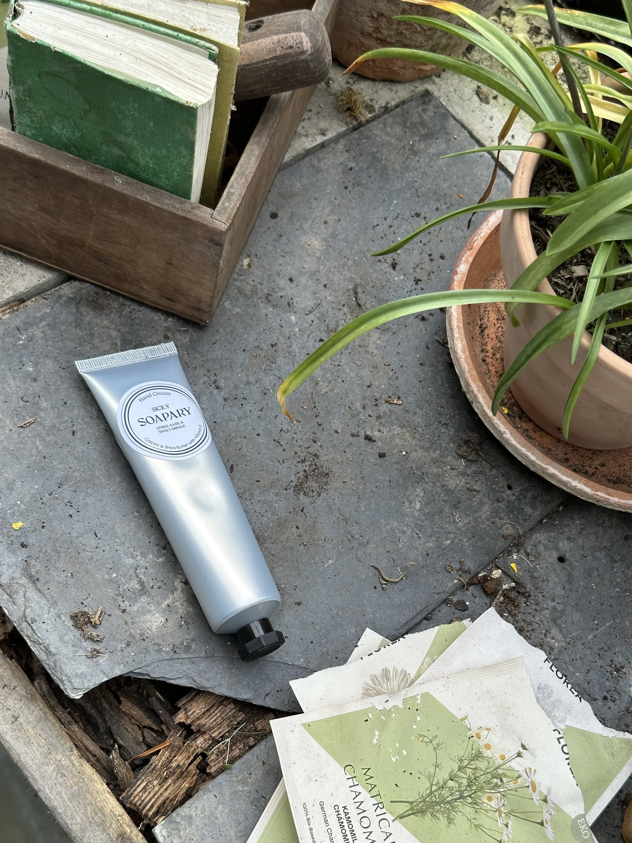 A wooden box containing green and white books, a tube of hand cream labeled Sicily Soapray, green leafy plants in clay pots, and several pamphlets with botanical illustrations and text on a concrete surface.