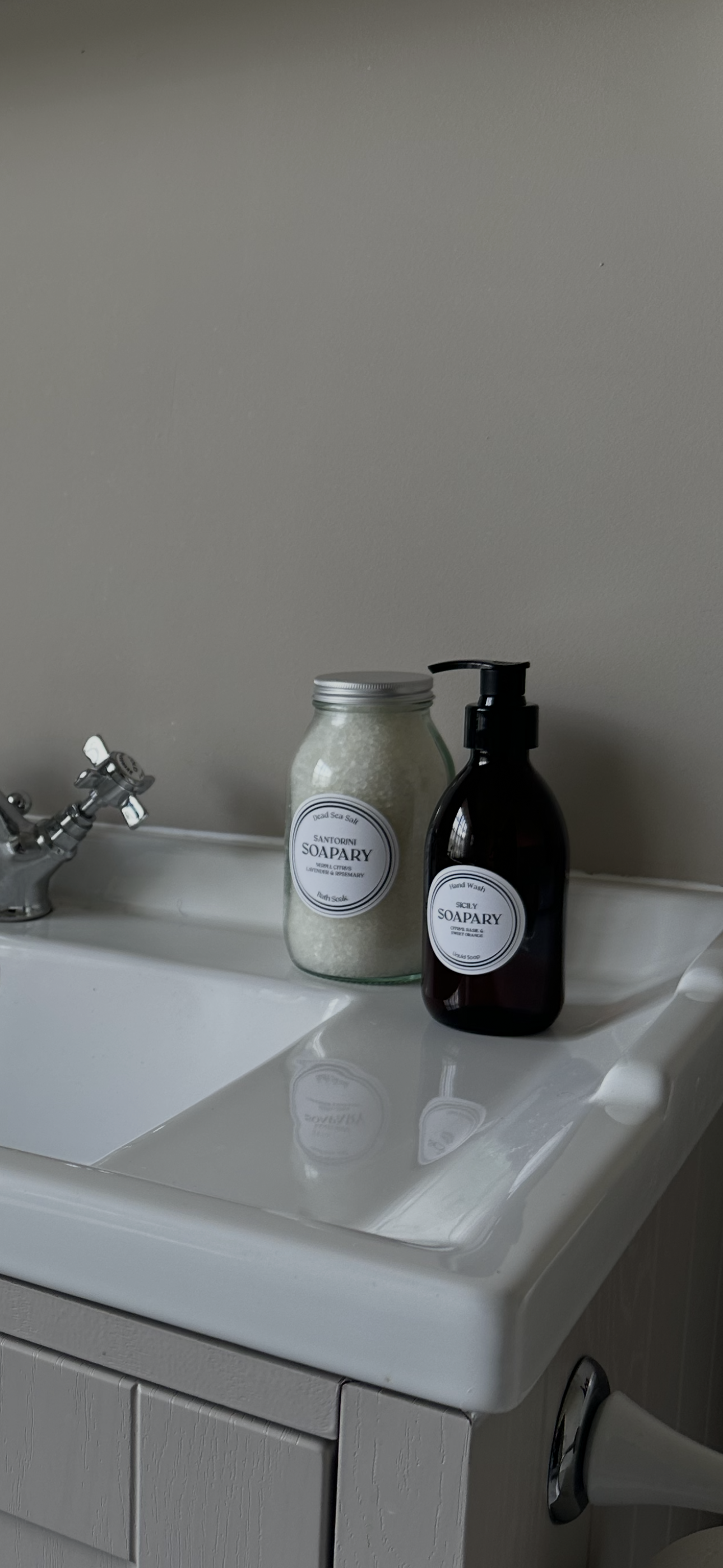 A white laundry sink with a silver faucet, holding a jar of Dead Sea salt labeled 'Santorini Soapary' and a black soap pump bottle also labeled 'Soapary.'
