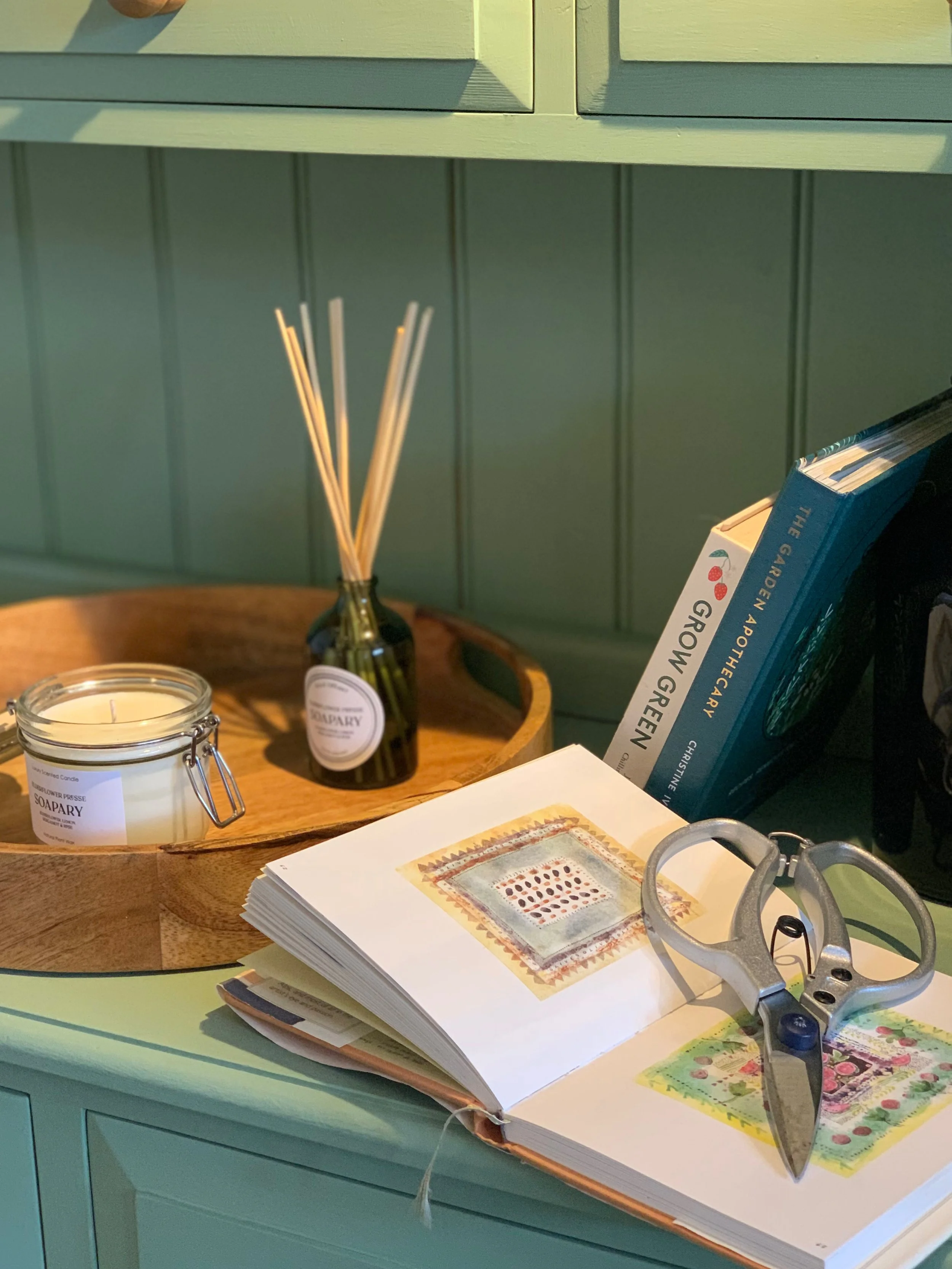 A tabletop with a glass jar with a candle, a black bottle with reed diffuser sticks, a stack of books including one titled 'Grow Green,' a pair of scissors, and an open art book with botanical illustrations.