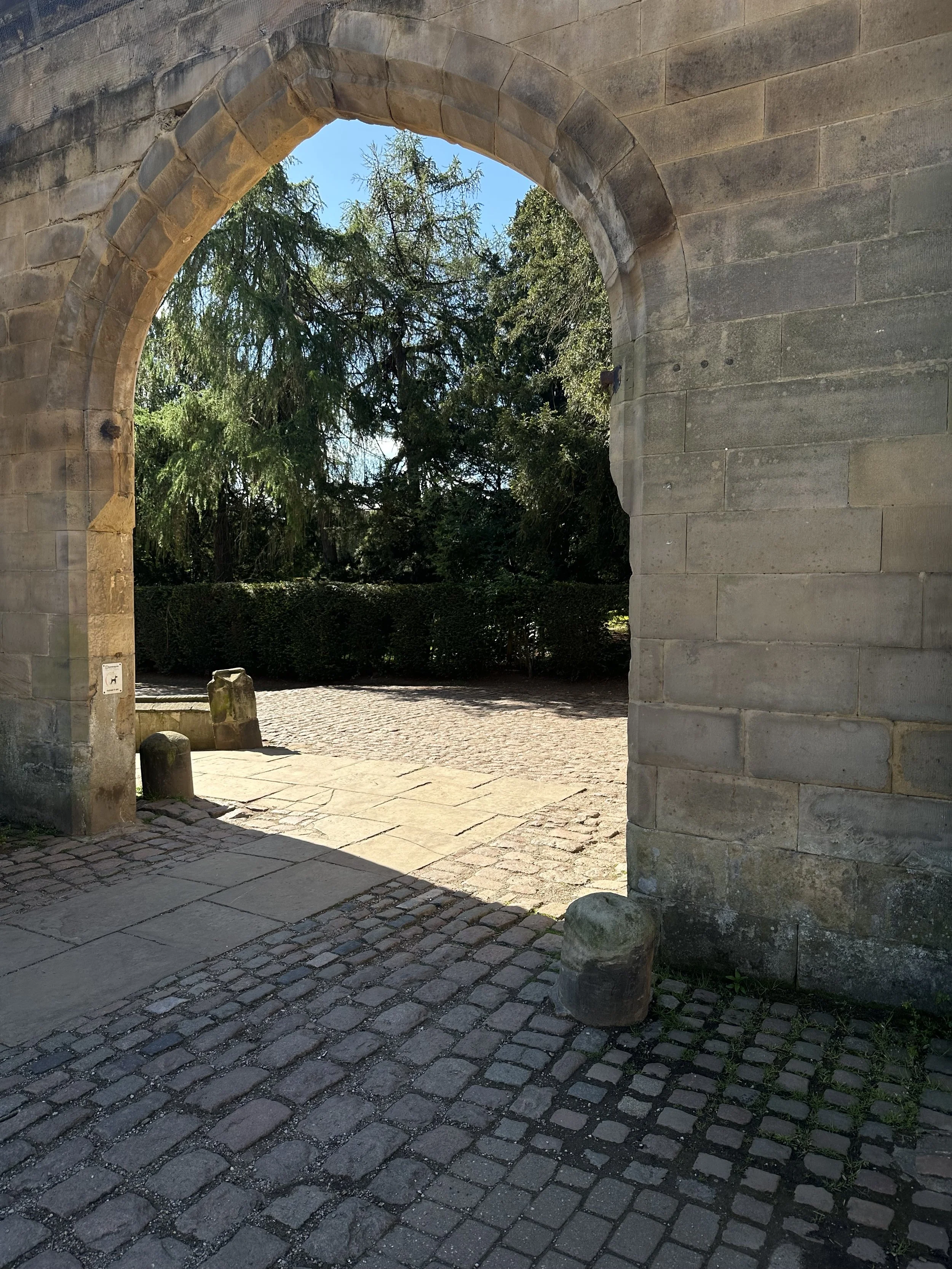 Stone archway opening to a sunny outdoor scene with trees and bushes, paving stones, and stone bollards.
