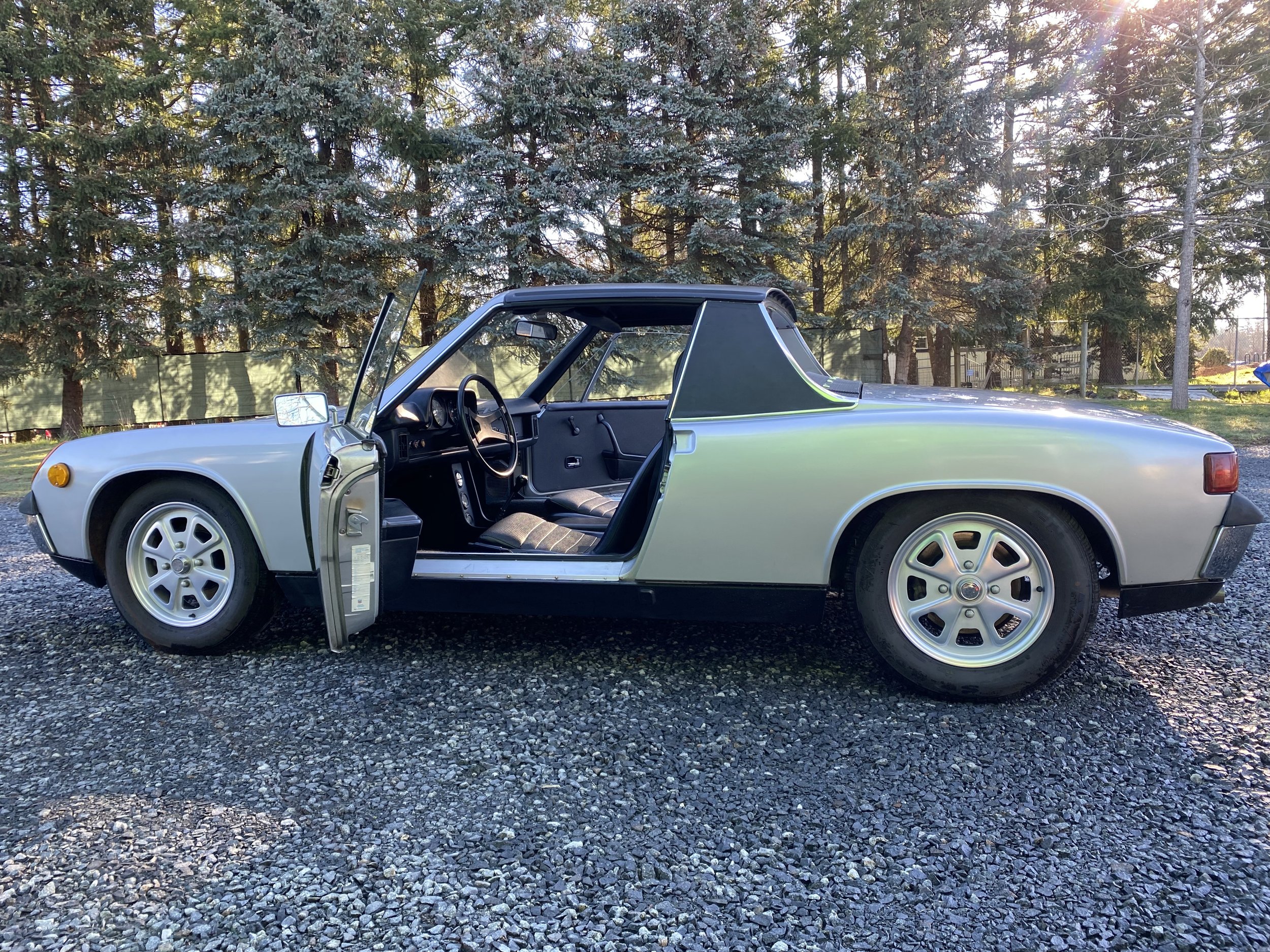 A vintage silver Porsche 914 car with the driver's side door open, parked on gravel with trees in the background.