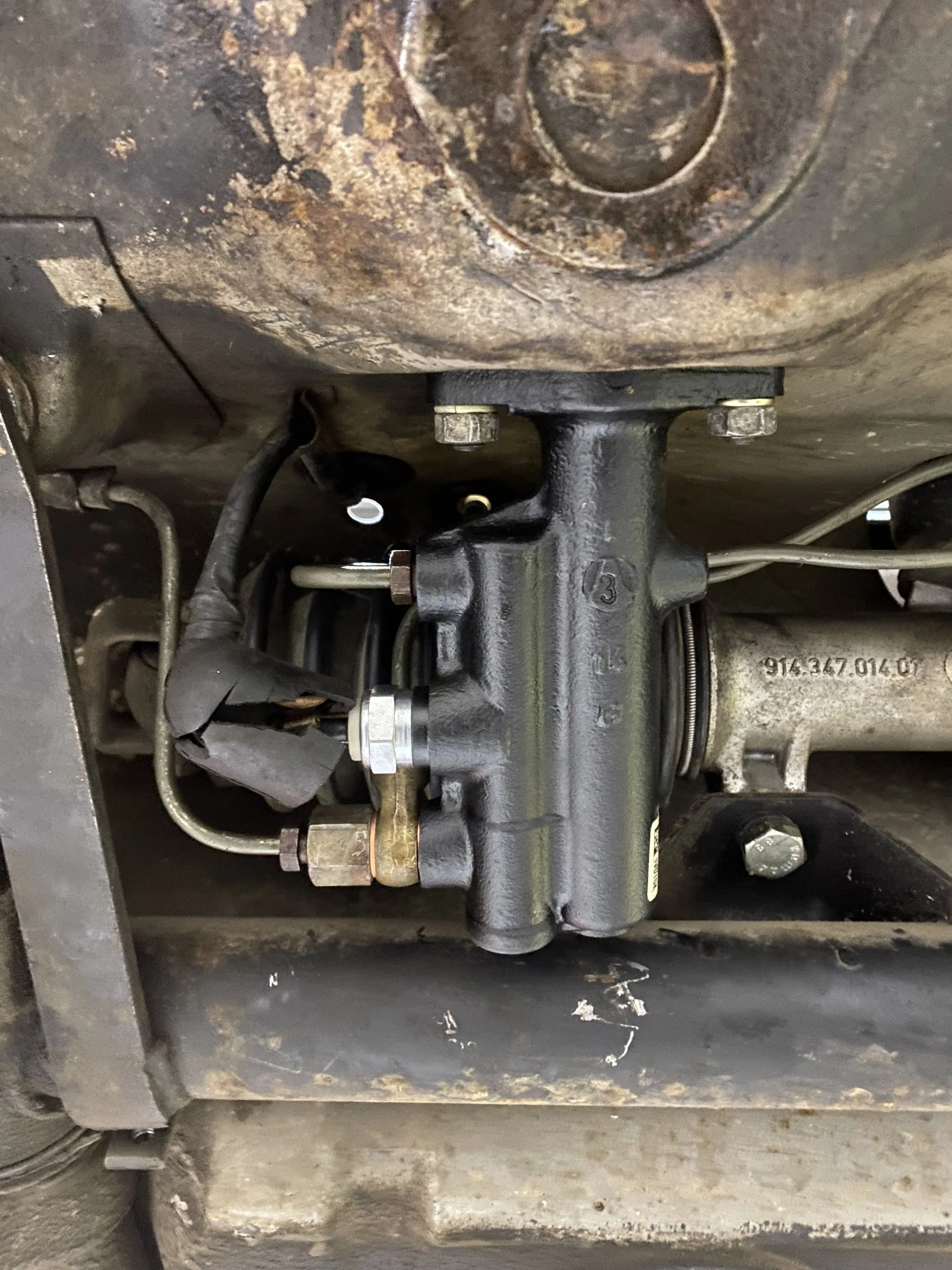 Close-up of a mechanical component from a 914, likely part of an engine or hydraulic system, featuring metal pipes, fittings, and a black cast metal part attached with bolts, with visible signs of rust and grime.