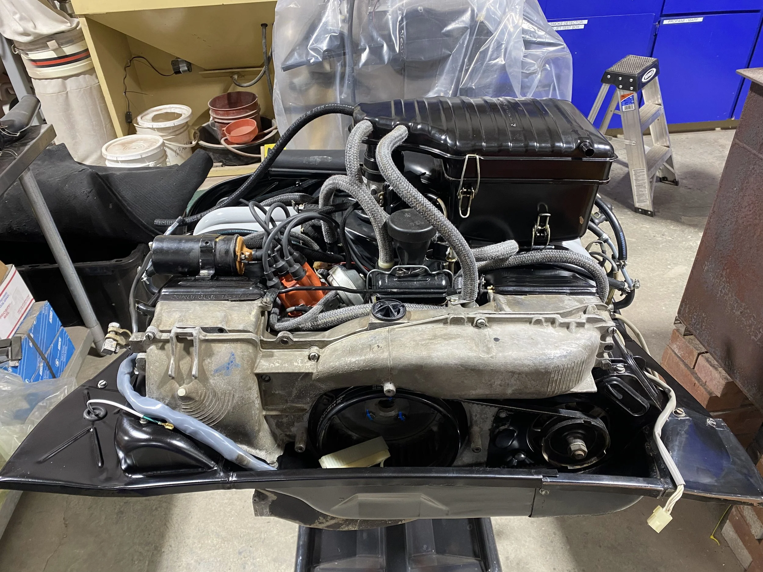 A Porsche 914 engine mounted on a workbench in a garage, surrounded by tools, parts, and storage cabinets.