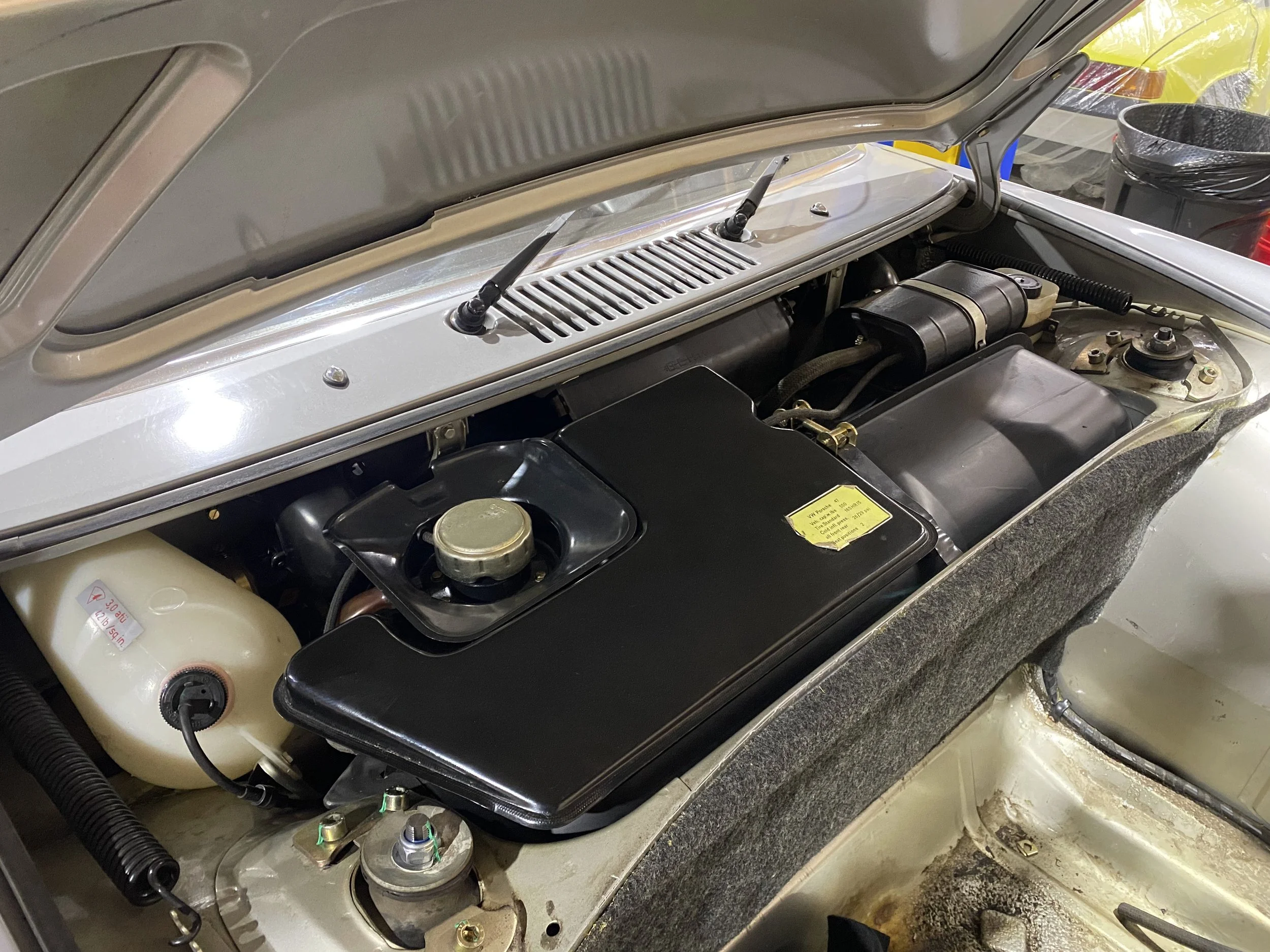Front compartment of a Porsche 914  with a black air filter cover, a coolant reservoir, and various engine components.