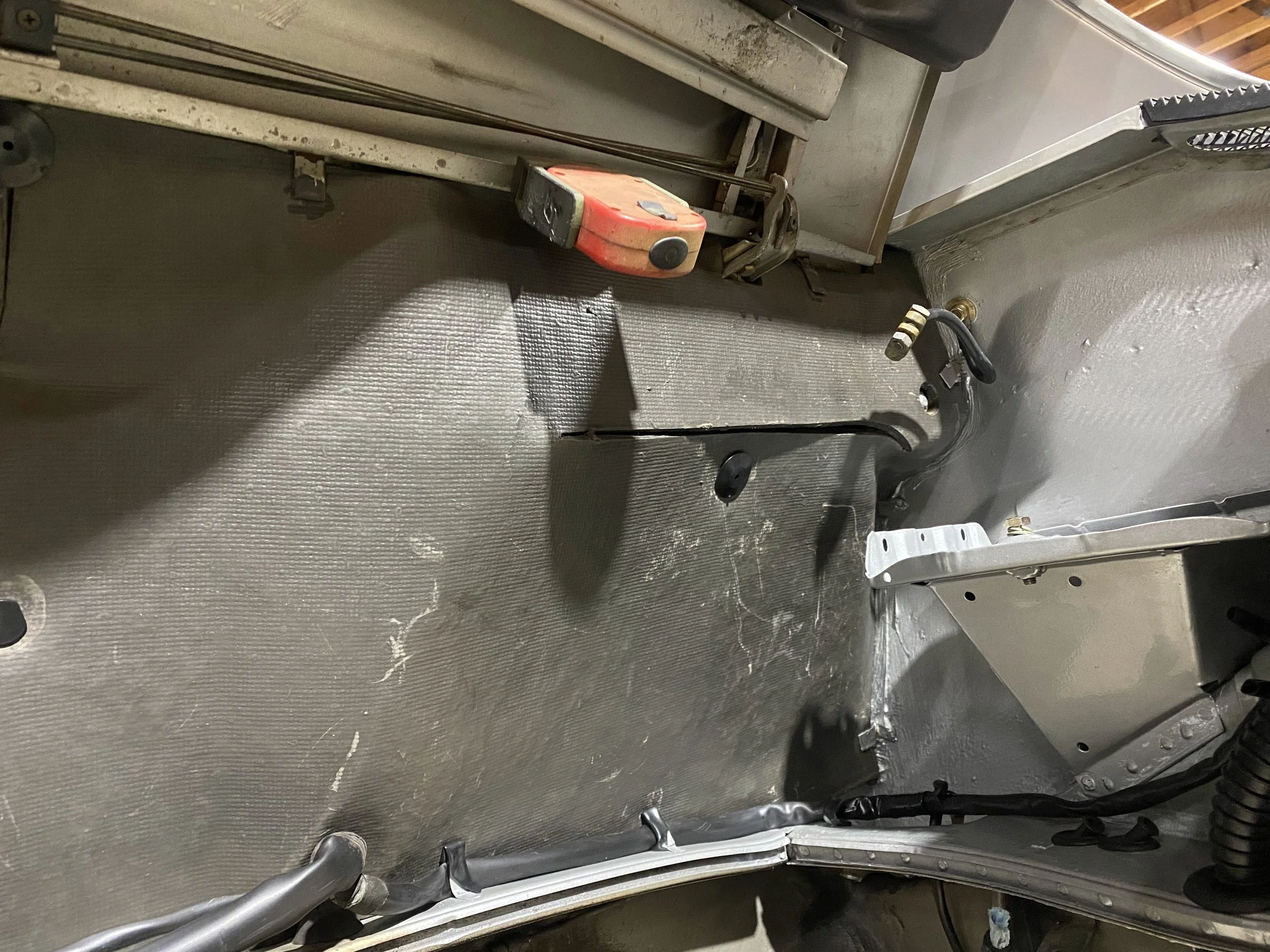 Interior of a Porsche 914 shows a textured metal floor with some dust and scratches, along with various wires, panels, and tools like a tape measure.