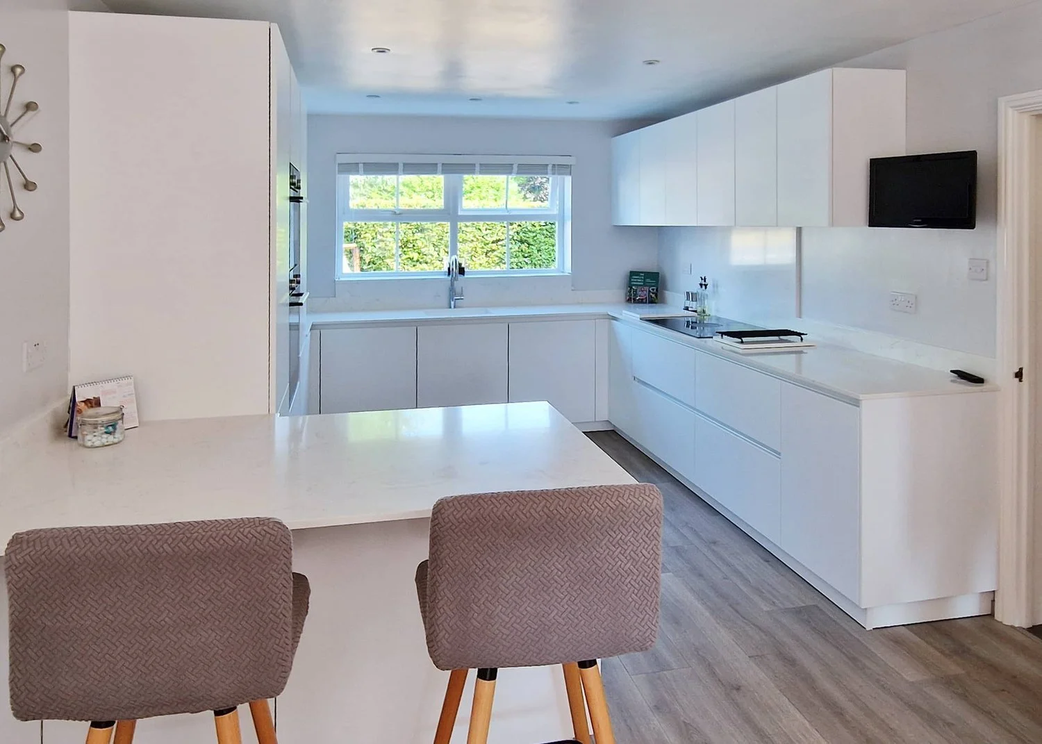 Modern white kitchen with large window, white cabinetry, built-in appliances, and a dining area with two beige chairs.