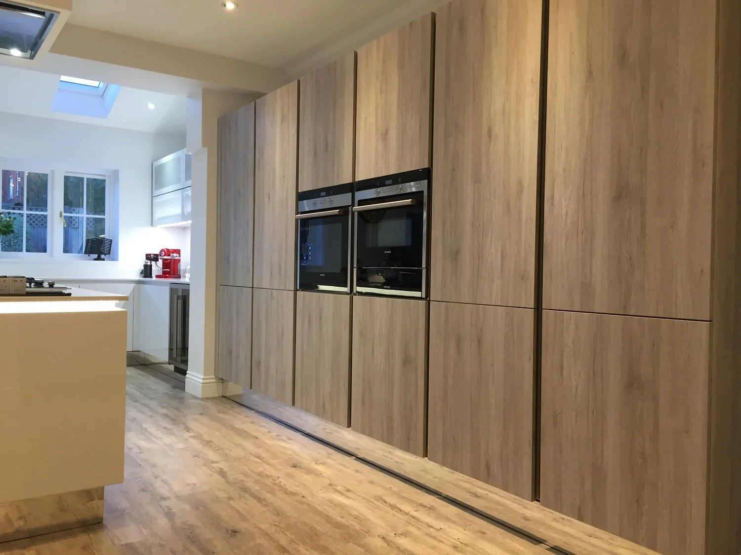 Modern kitchen with wood-paneled cabinets, built-in double oven, and a section of light-colored countertop with a red espresso machine near a window.