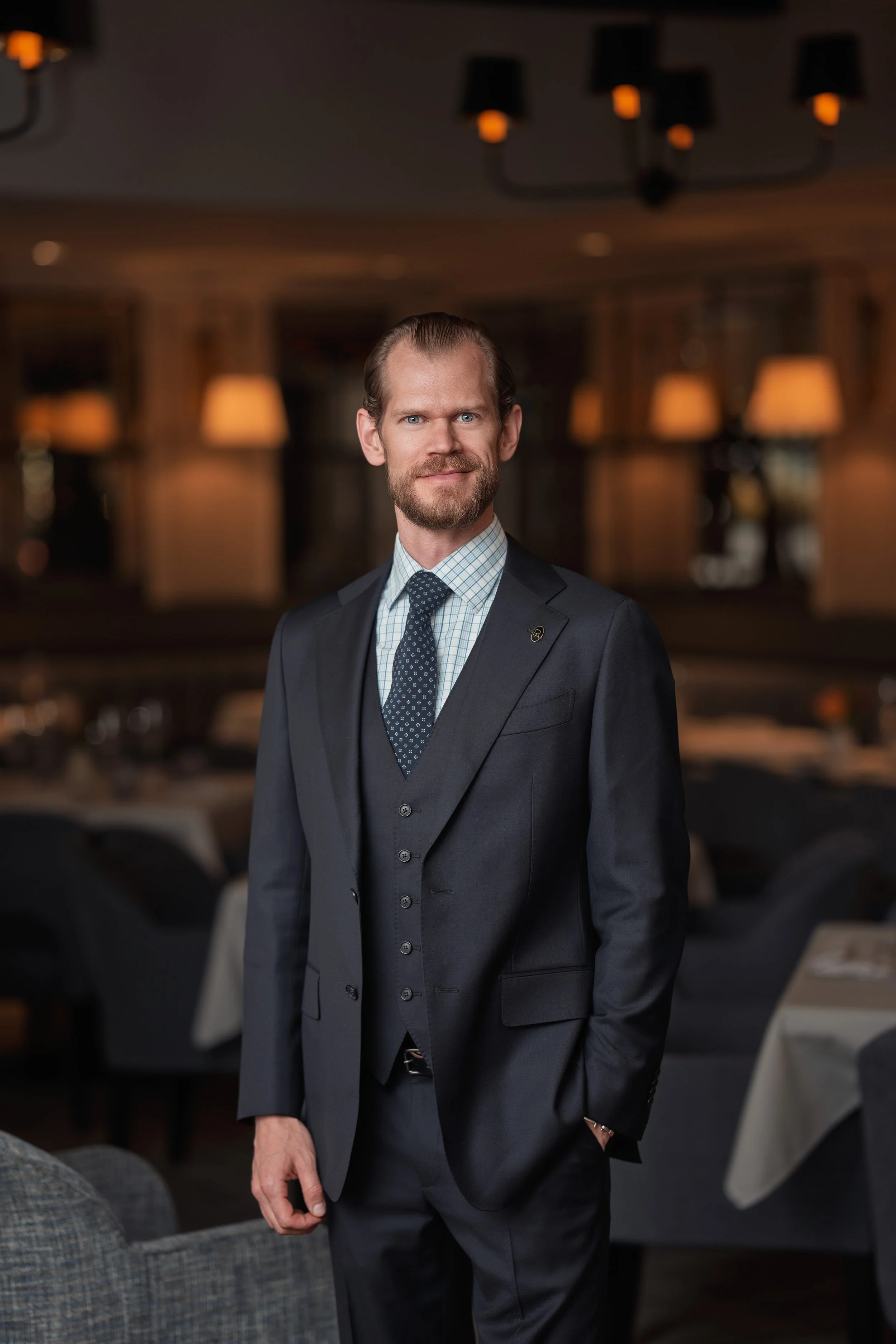 A man with a beard and slicked-back hair wearing a dark suit and patterned tie standing in a restaurant or banquet hall with dim lighting and round tables in the background.
