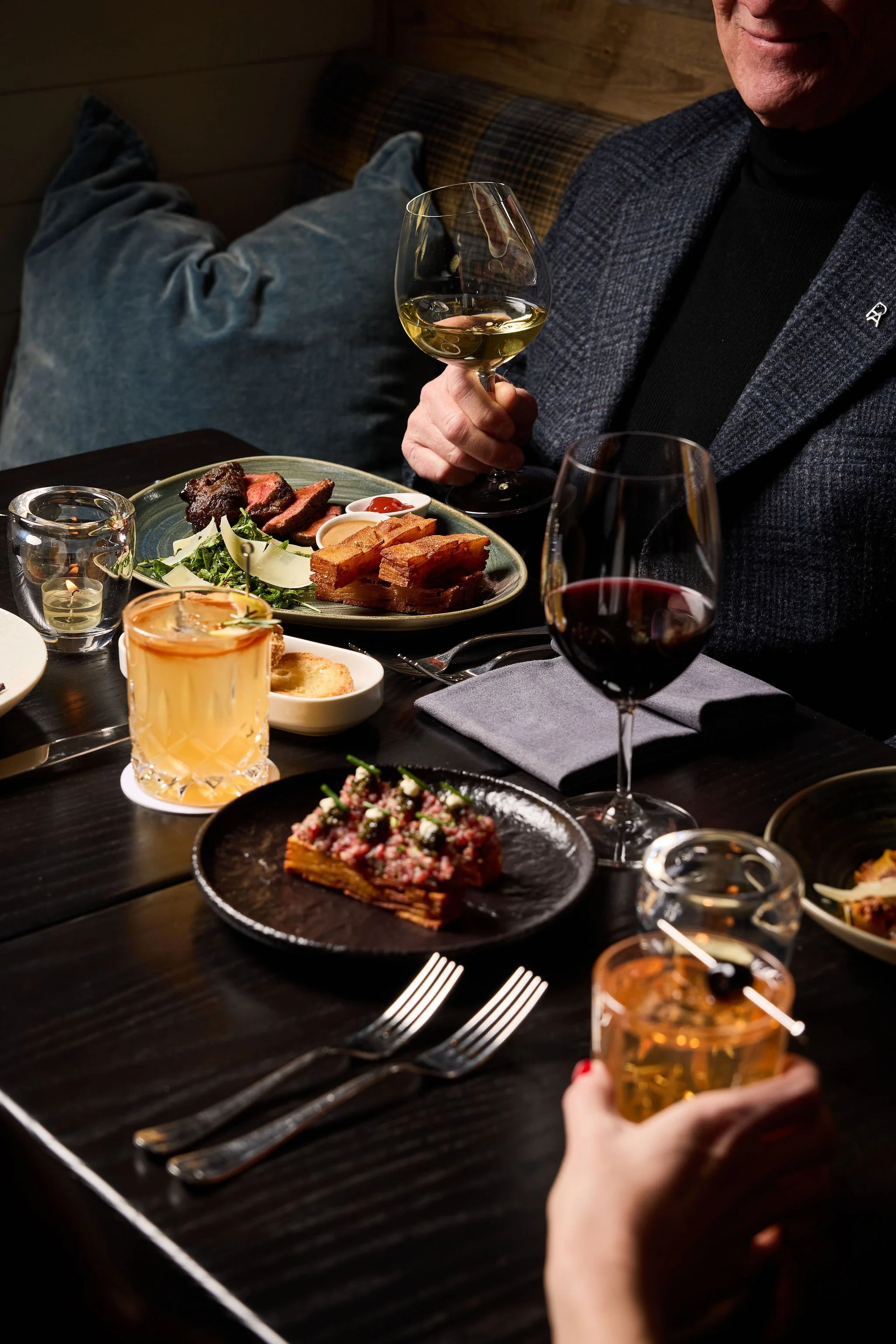 A person in a dark blazer holding a glass of wine at a dinner table with various dishes, including steak, salad, and dessert, in a cozy restaurant setting.