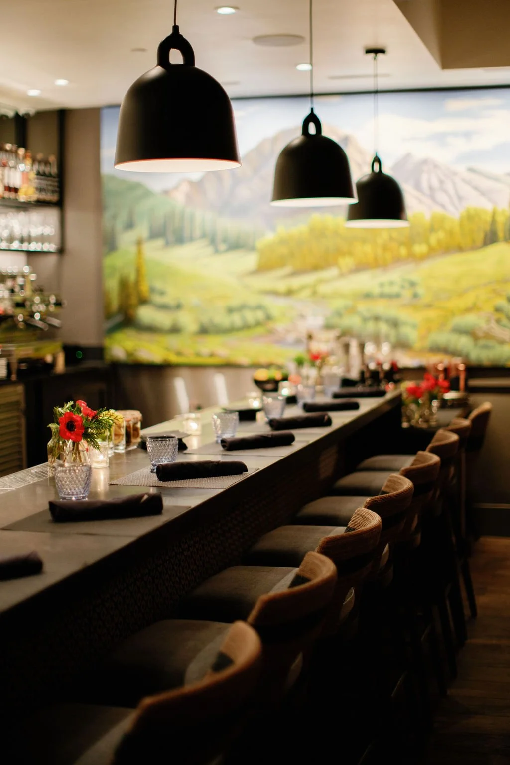 Interior of a restaurant with a long counter, decorated with small flower arrangements, pendant lights, and a mural of a landscape with mountains and trees.