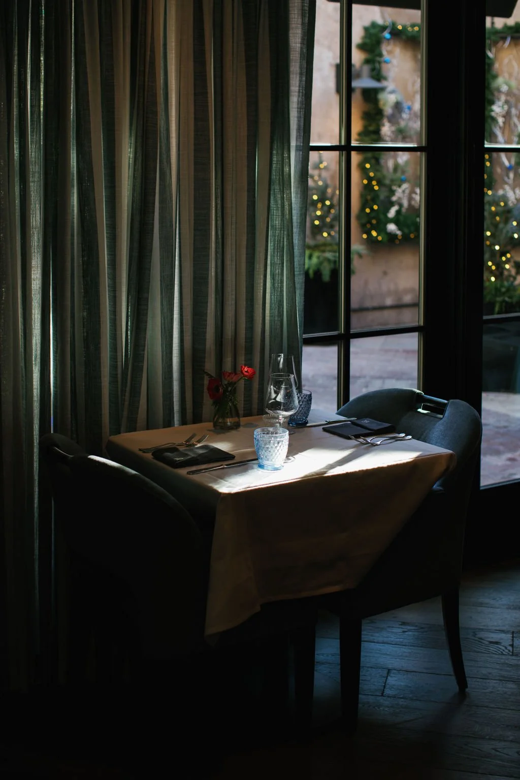 Window Table for Two seat for a romantic dinner with a slate blue curtain. The sun is peeking in through the windows creating a ray of light on the table.