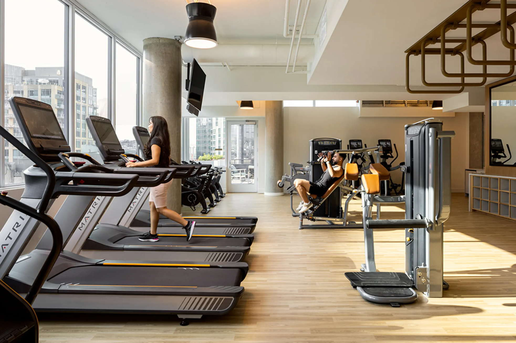 Indoor gym with large windows, showing two women on treadmills and various exercise machines, with city buildings visible outside.