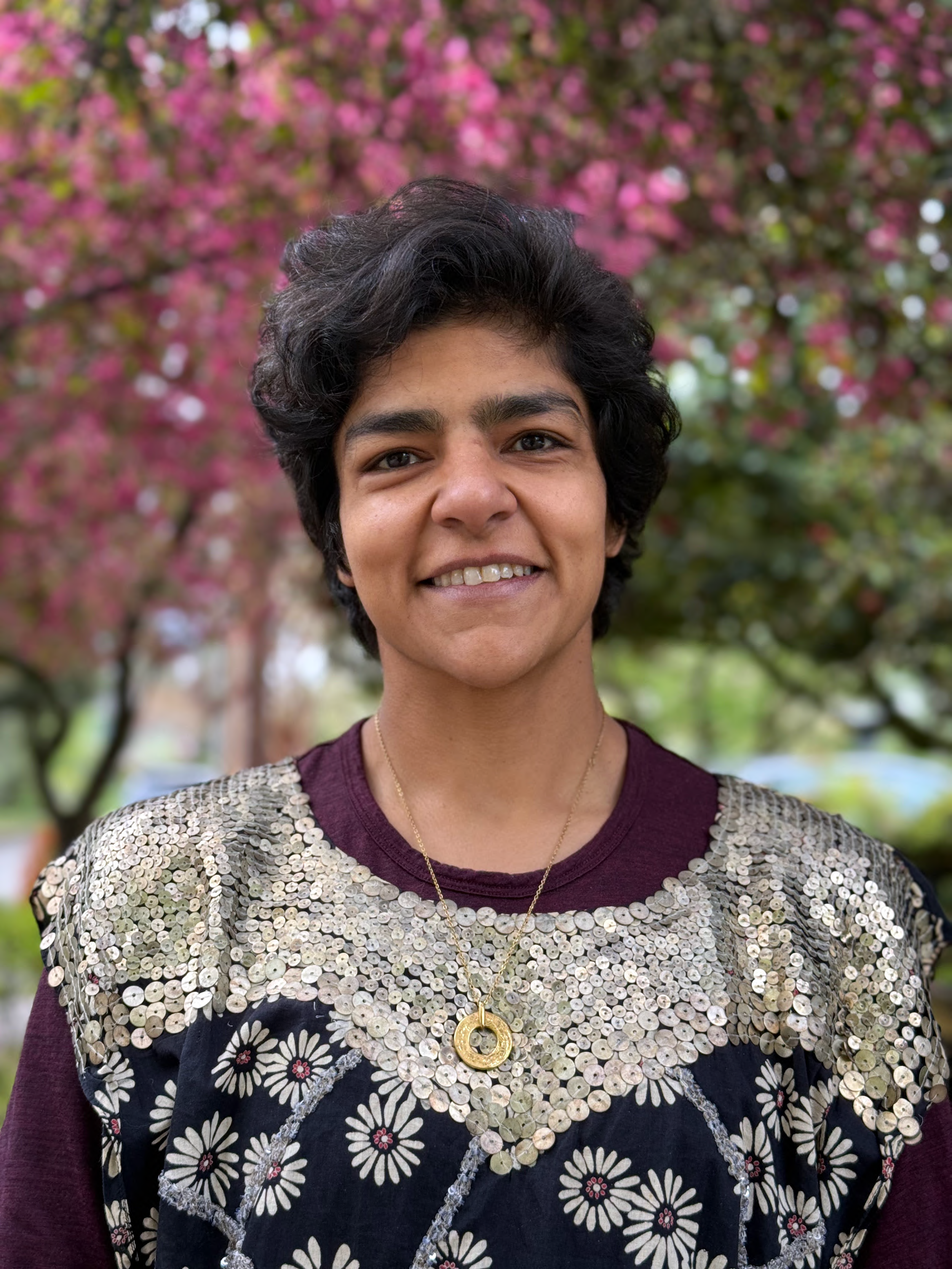 A woman with short, curly black hair smiling outdoors in front of a blooming pink-flowered tree, wearing a sequined and floral patterned black and maroon top with a gold necklace.