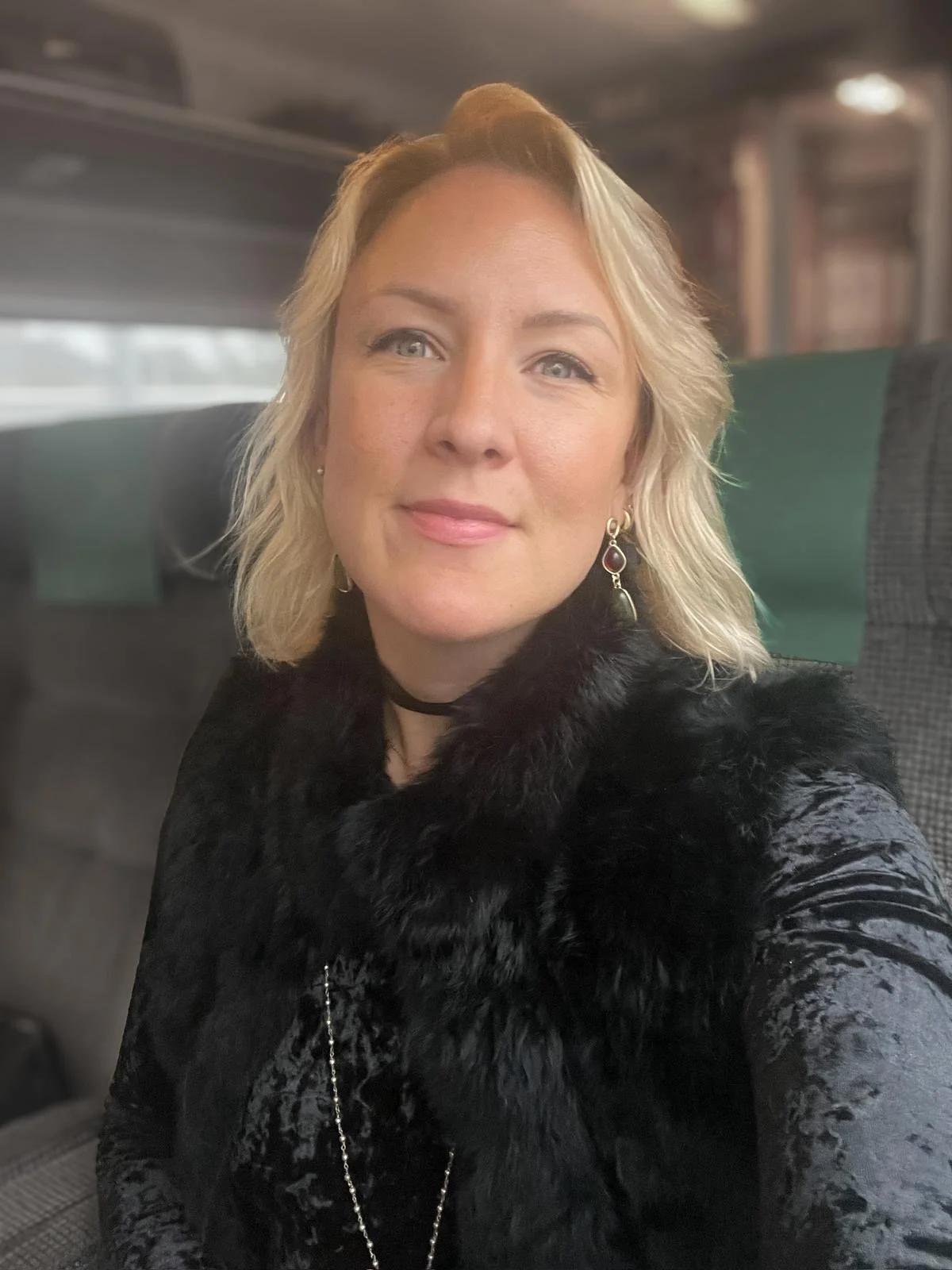 A woman with blonde hair, blue eyes, and light skin smiling on a bus. She's wearing earrings, a black fur coat, and a black choker.