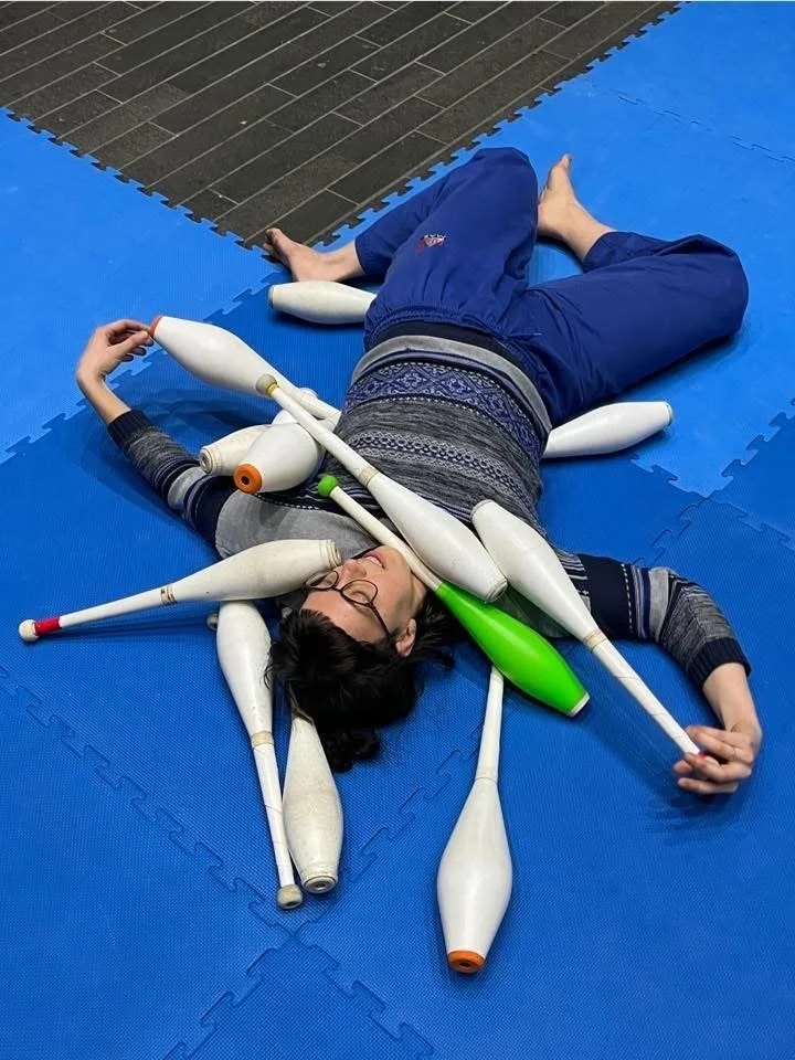 Person lying on a blue mat surrounded by juggling clubs, with some clubs resting on their body and the floor.