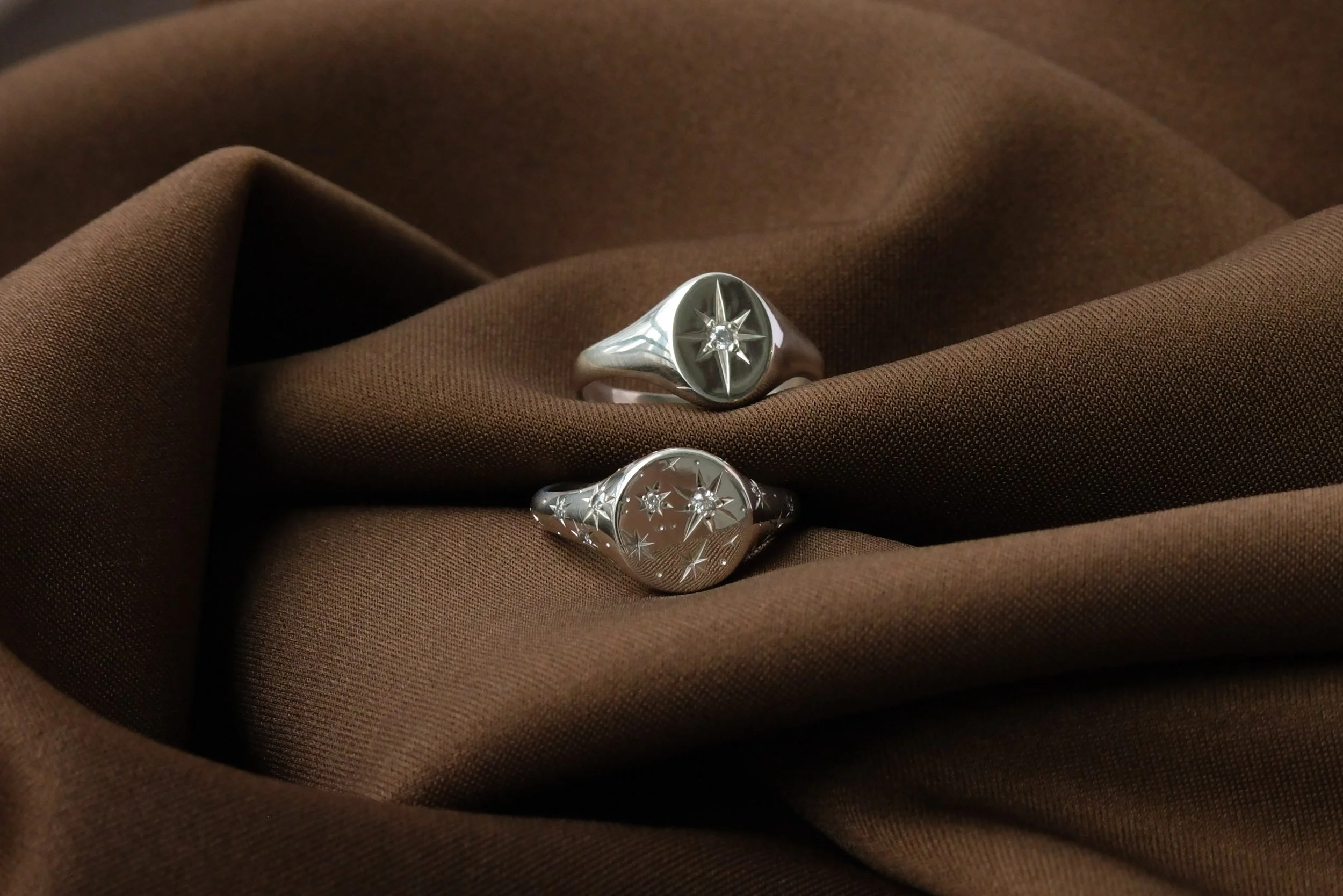 Two silver rings with star-shaped engravings and small gemstones, displayed on brown fabric.