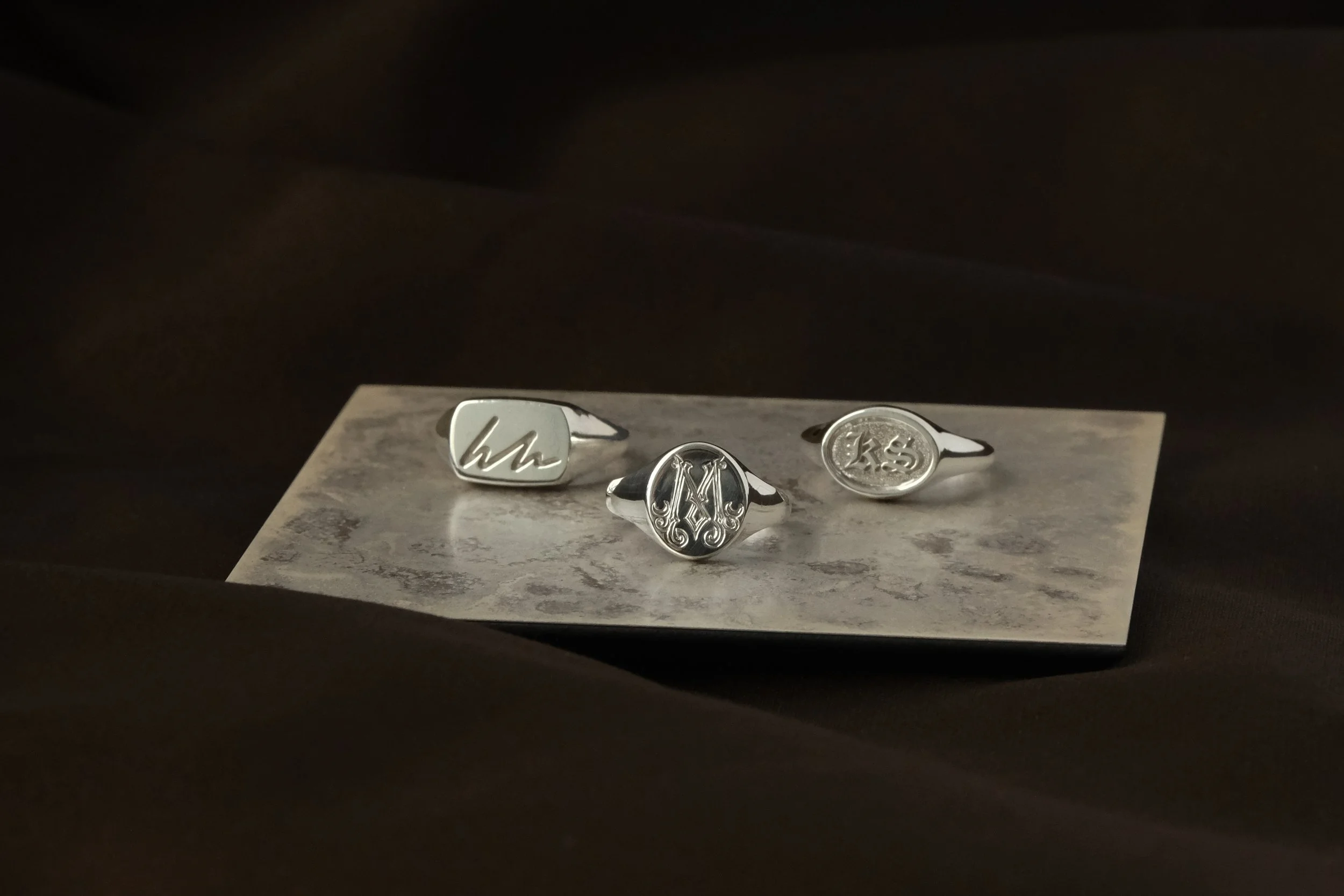 Three silver rings with engraved initials, displayed on a textured surface within a black box.