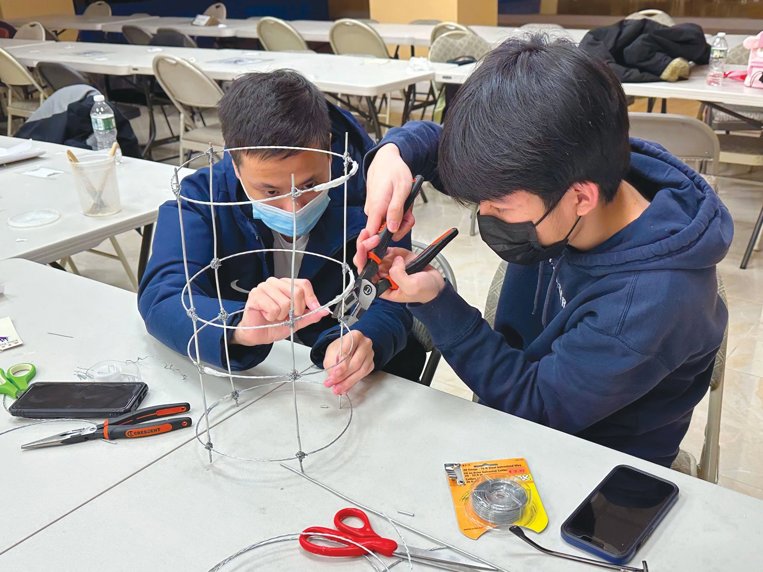 Two students engage in mentorship while using hand tools to build a wire sculpture in a collaborative workshop.