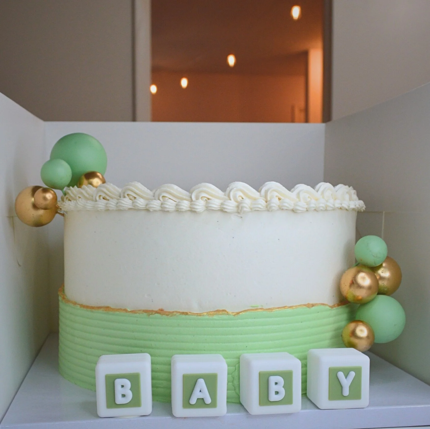 #126. This is a 10" round, 3-layer baby shower cake with a ribbed frosting overlay, edible gold paint over the top edge of the overlay, and cake ball and "BABY" block decor.