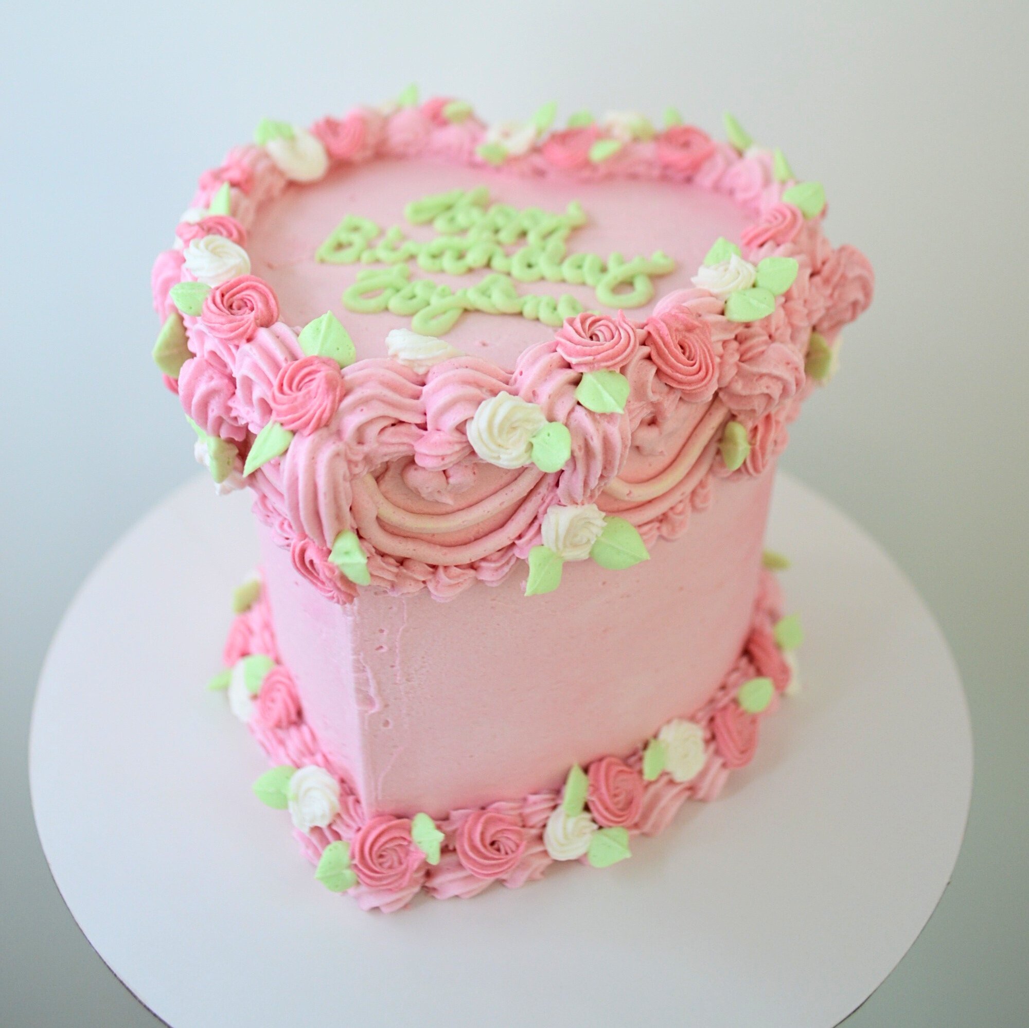 #33. Pink, heart-shaped vintage style cake with whit and pink rosettes, green leaves and a custom message.
