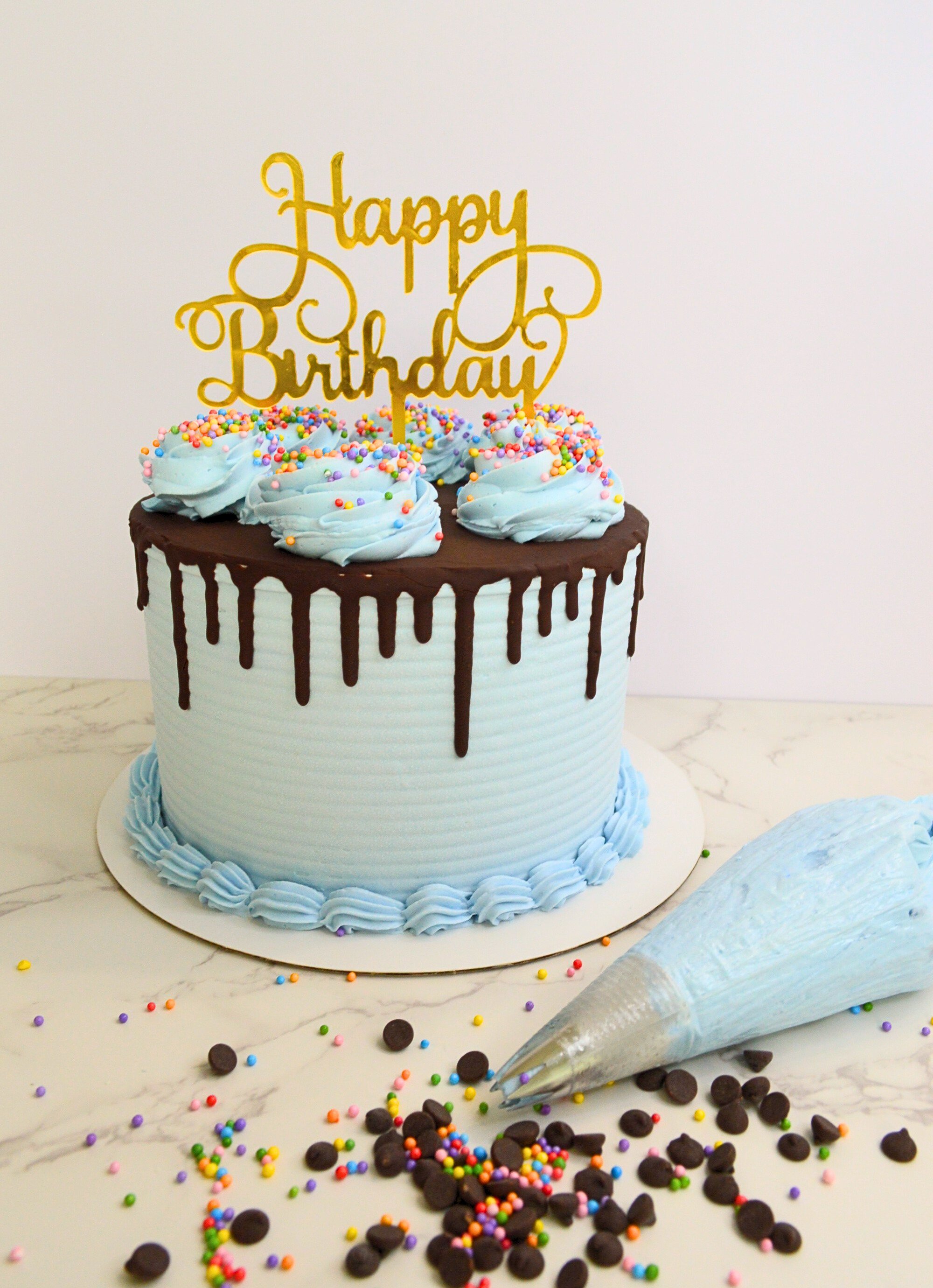 #71. Blue drip cake with a bottom border, swirls, rainbow sprinkles and a gold "Happy Birthday" topper.
