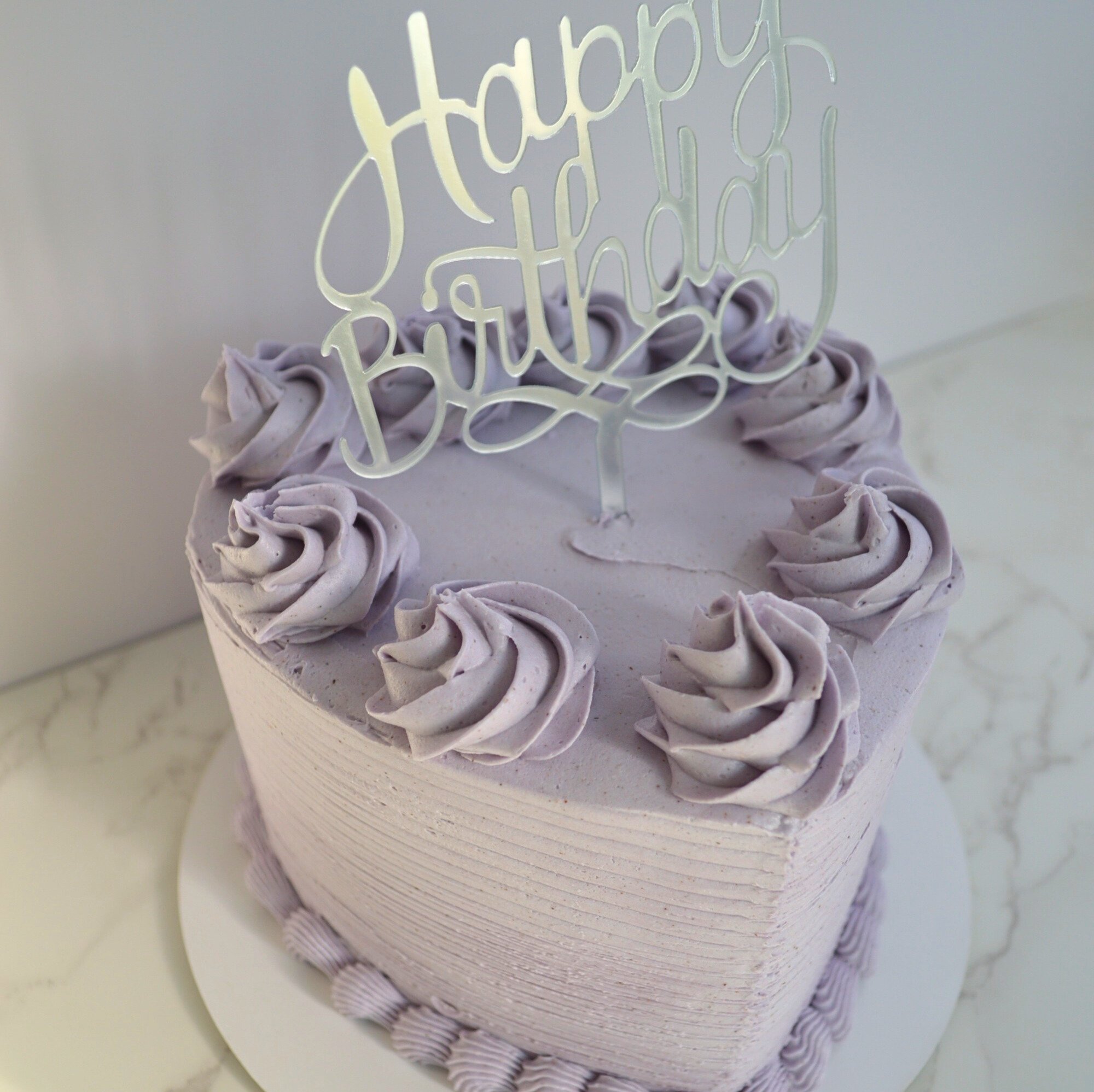 #37. Dusty purple, heart-shaped cake with a ridged outer texture, bottom border, swirls on top and a silver "Happy Birthday" topper.