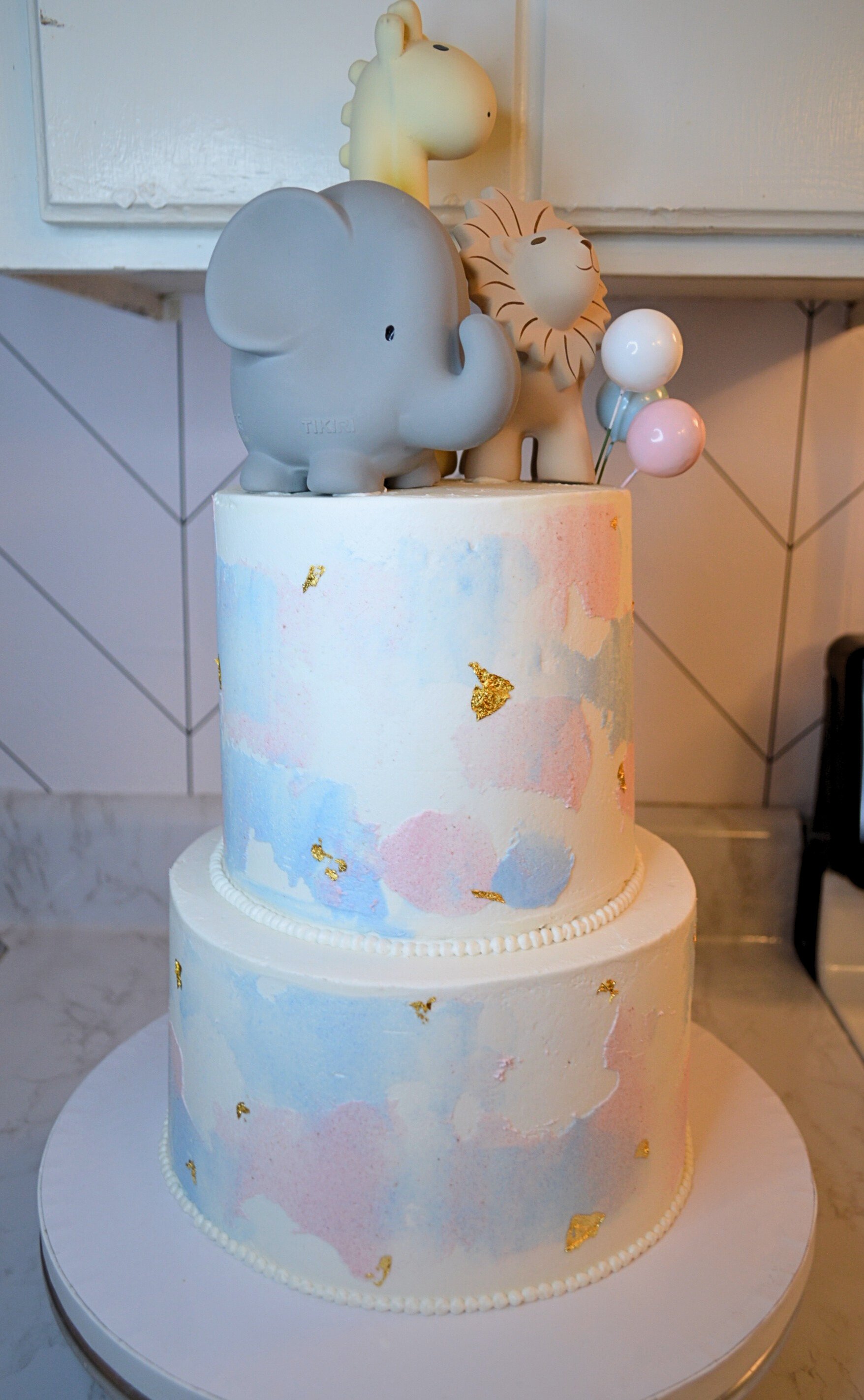 #22. Two-tiered baby shower cake with pink and blue frosting splotches smoothed onto the cake, edible gold foil, and custom decor.
