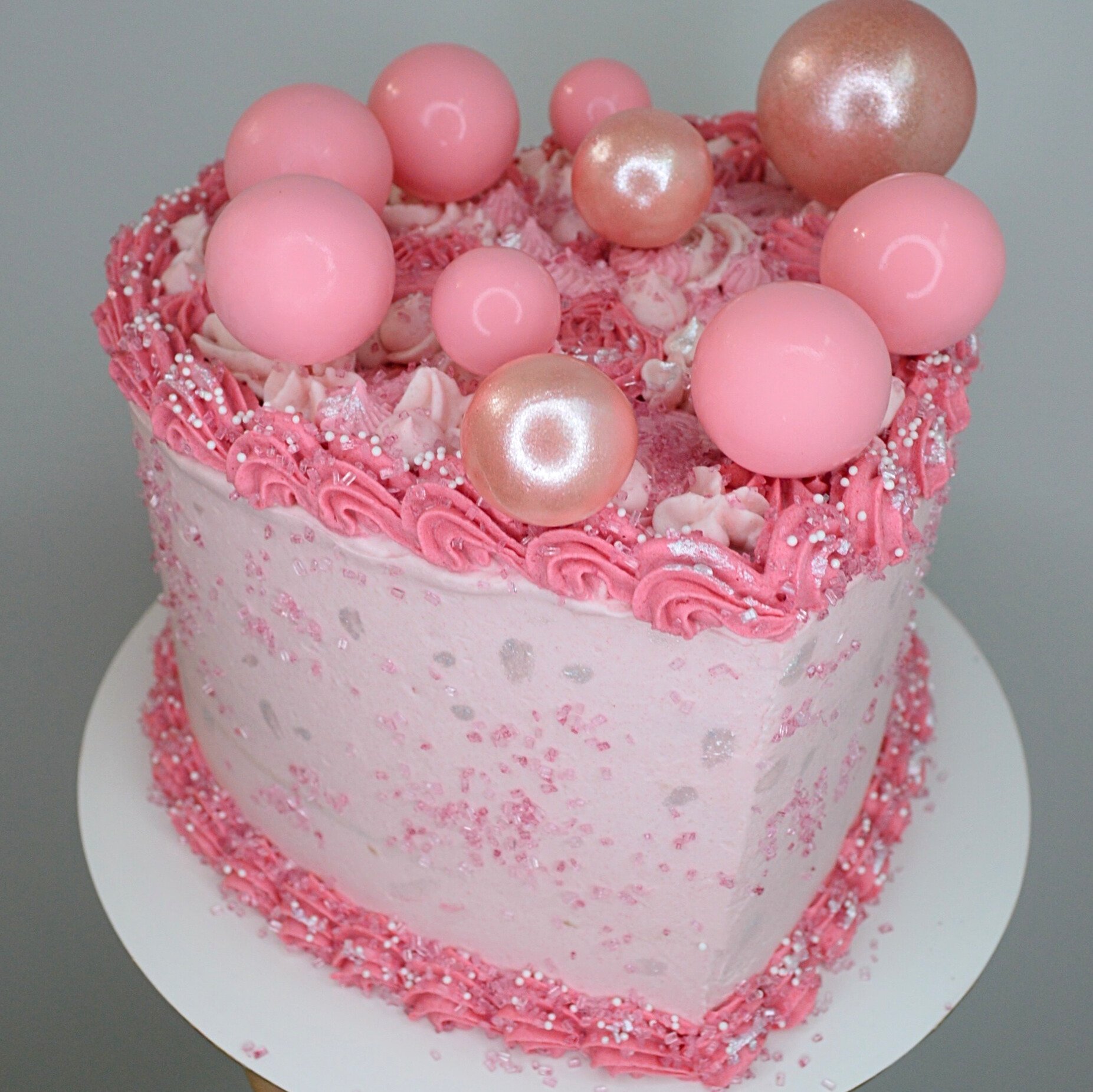 #13. Pink heart-shaped cake with a darker pink top and bottom border, pink sugar sprinkles and decorative cake balls.