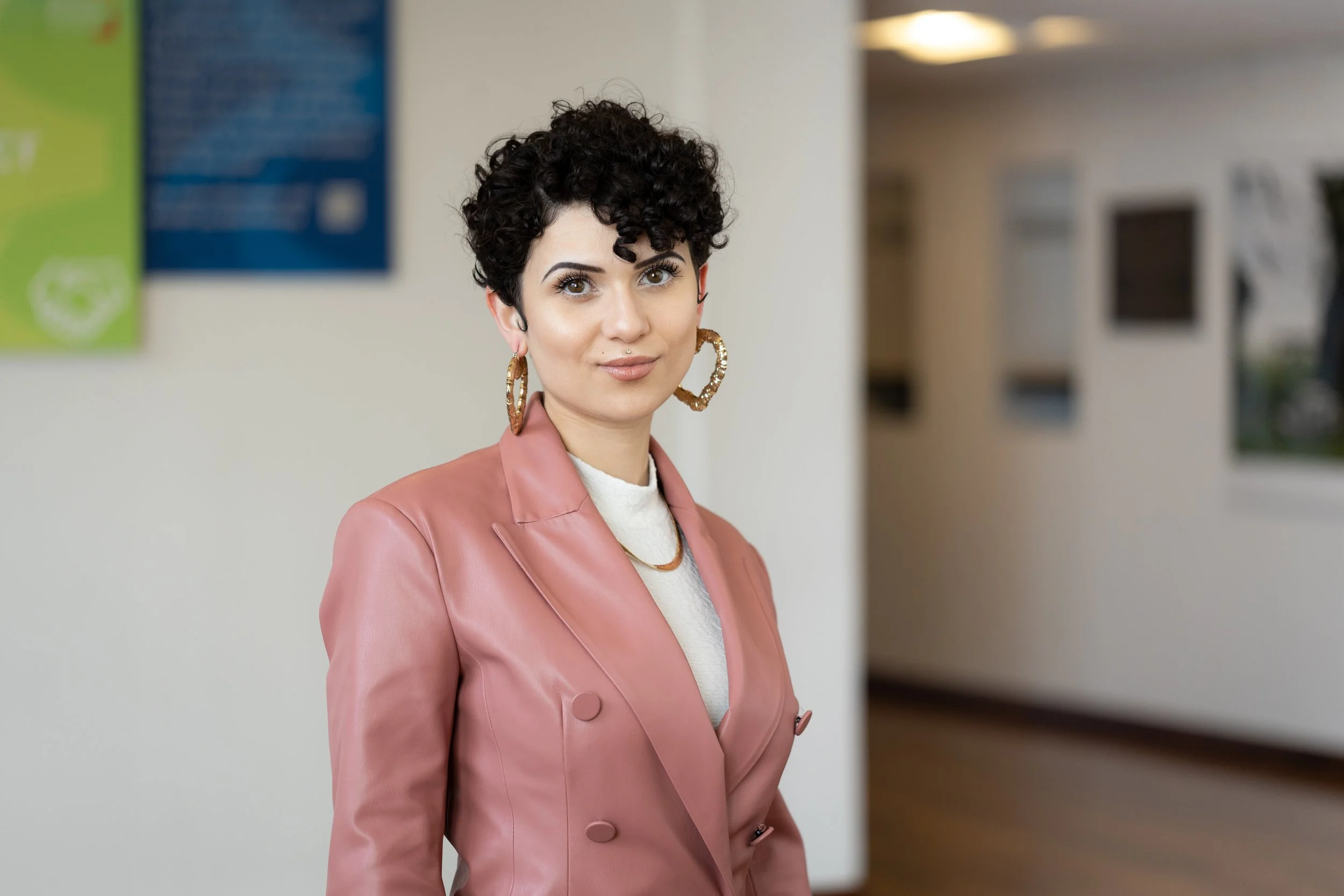 A woman with short curly black hair and hoop earrings standing indoors, wearing a pink blazer over a white top.