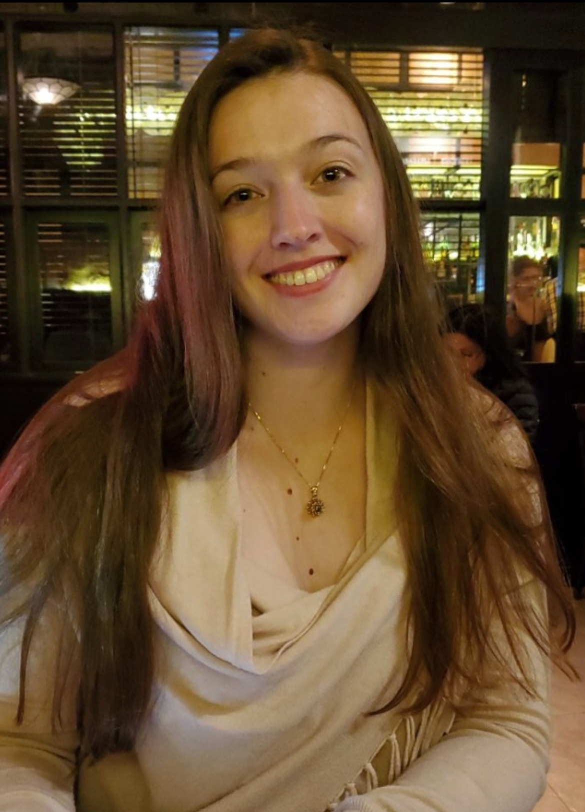 A woman with long brown hair smiling, sitting indoors at a restaurant or cafe with a warmly lit background.