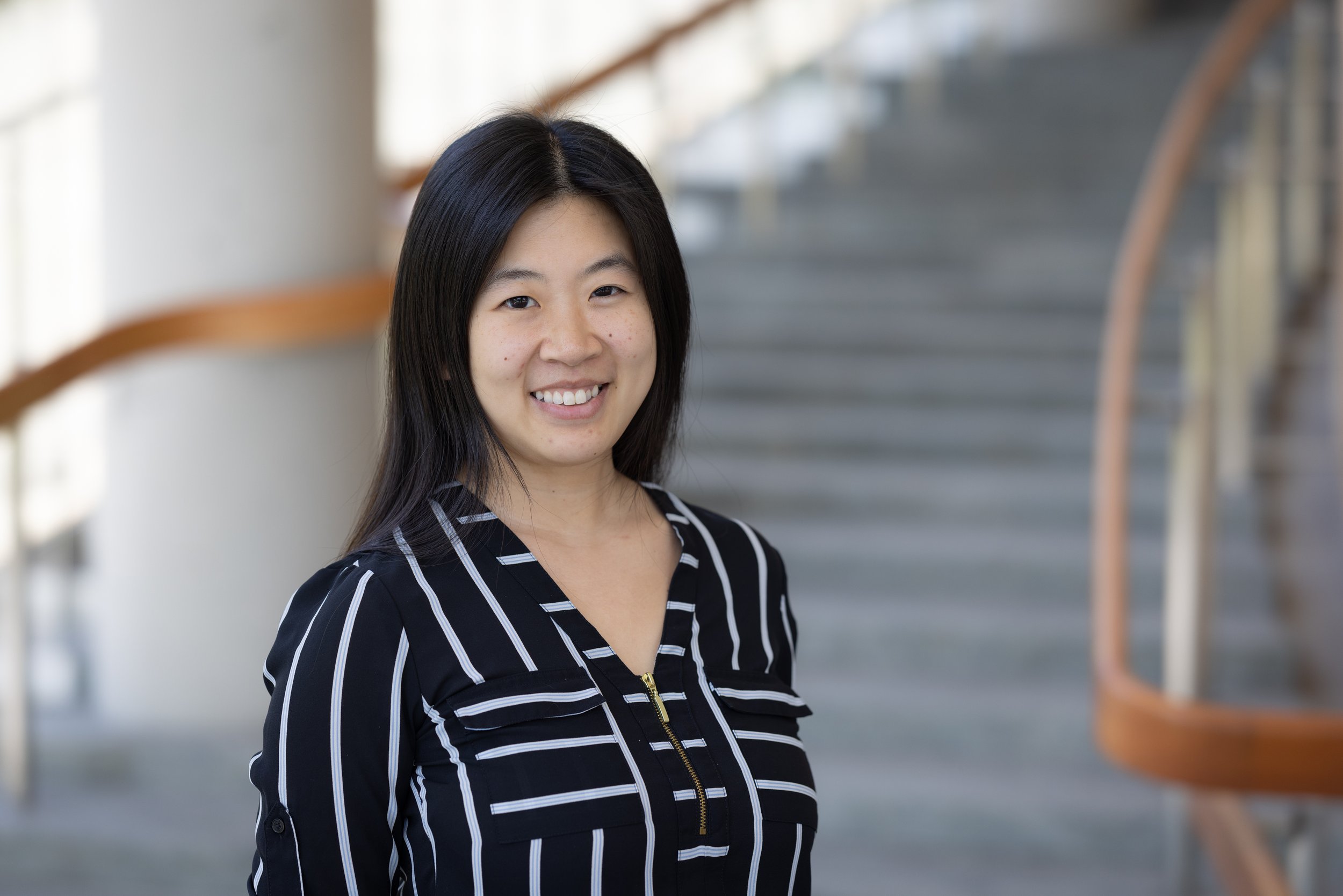 A young Asian woman with black hair smiling, wearing a black and white striped shirt, standing indoors near a staircase with wooden handrails.