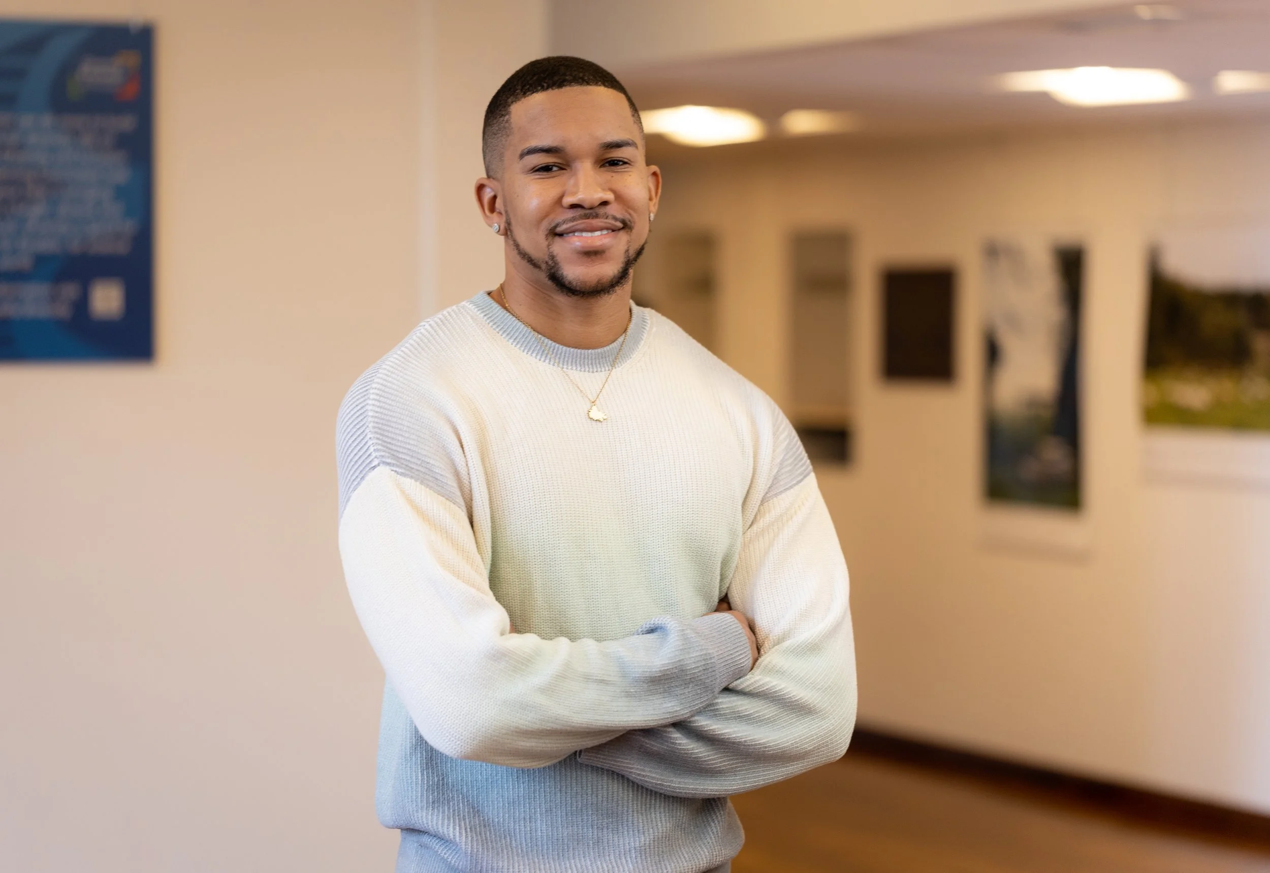 A young man with short hair and a beard, wearing a light-colored sweater and a gold necklace, standing with arms crossed and smiling in an indoor setting with artwork on the walls.