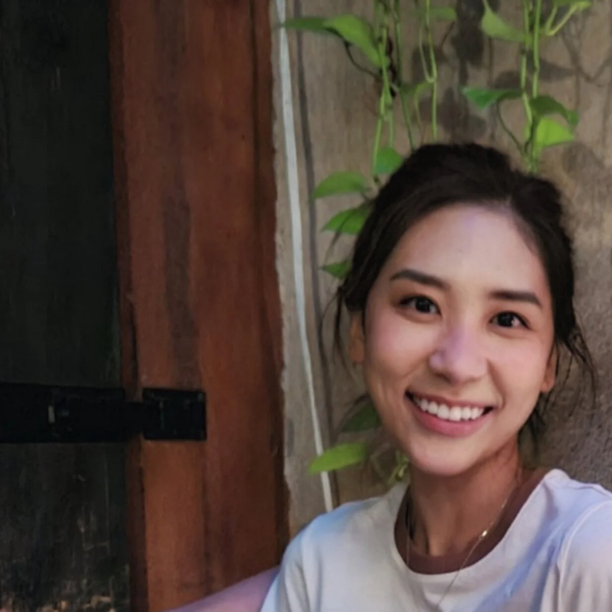 A smiling young woman with dark hair tied back, wearing a white top, standing near a wooden door and green leafy vines.