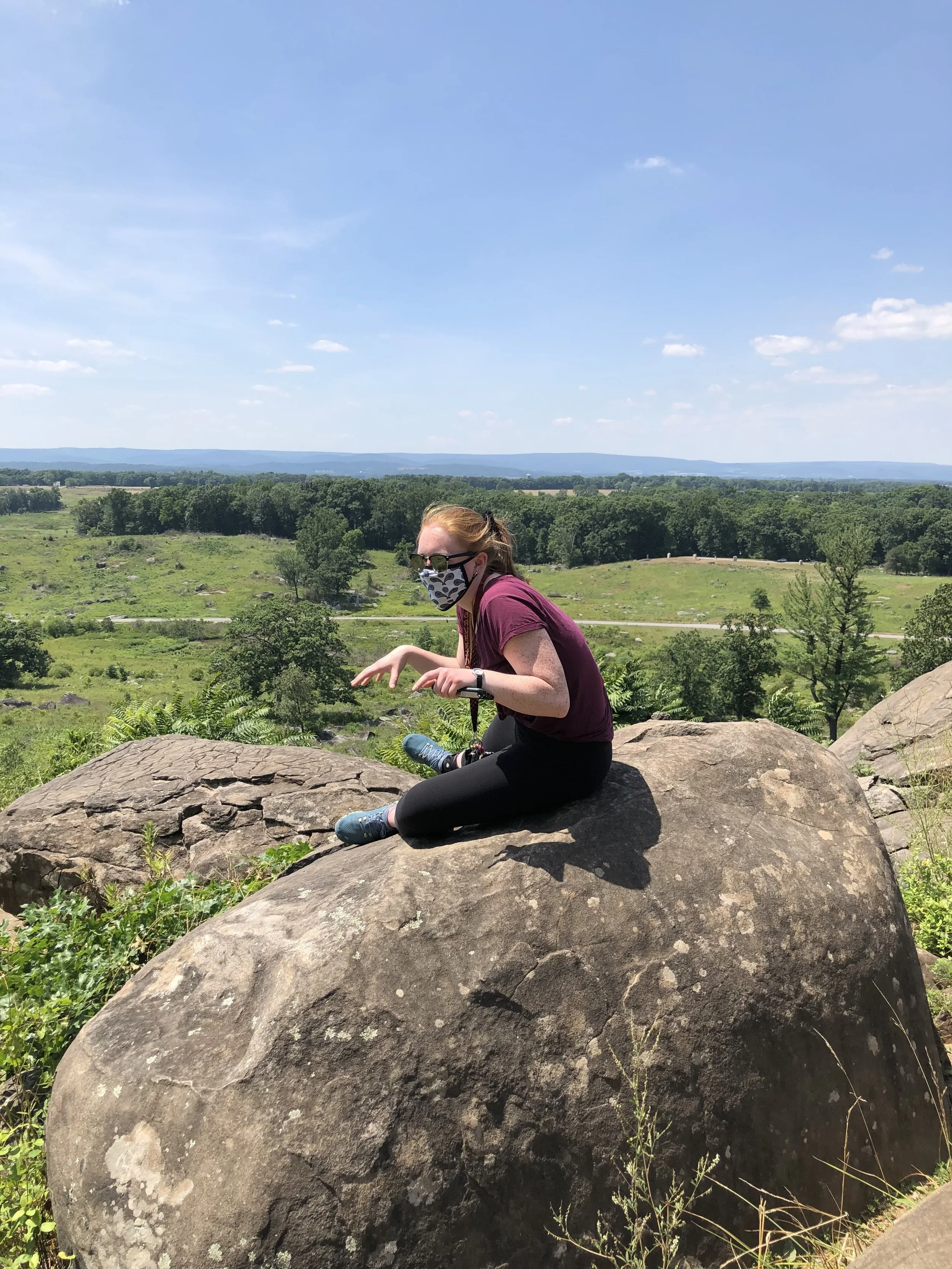 A woman sitting on a large rock outdoors in a scenic, green landscape under a blue sky, wearing a face mask, sunglasses, and casual clothing.
