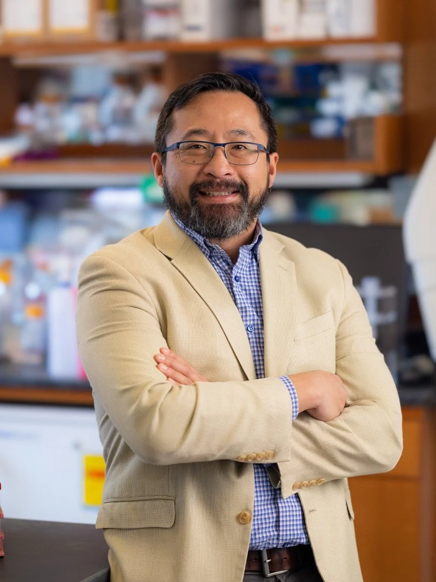 A man with glasses and a beard smiling with arms crossed, standing in a laboratory or pharmacy with shelves of supplies behind him.