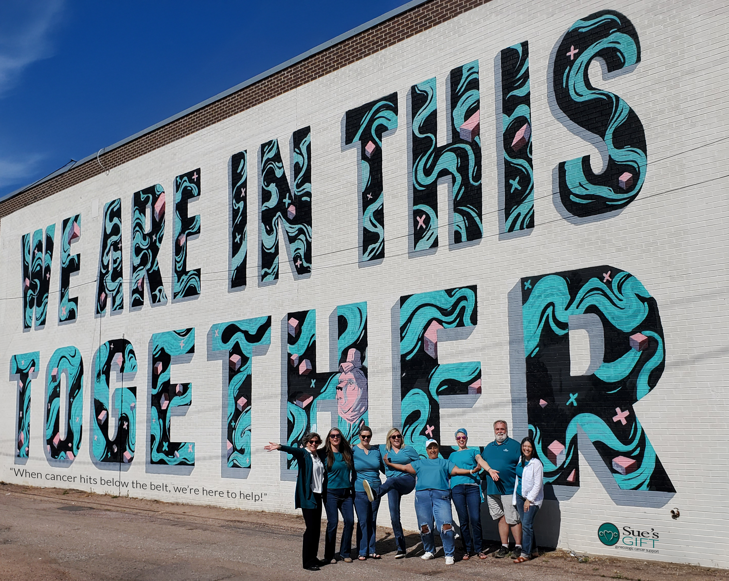 Sue's Gift Board of Directors standing in front of a giant teal and black lettering mural that reads, "We're In This Together"