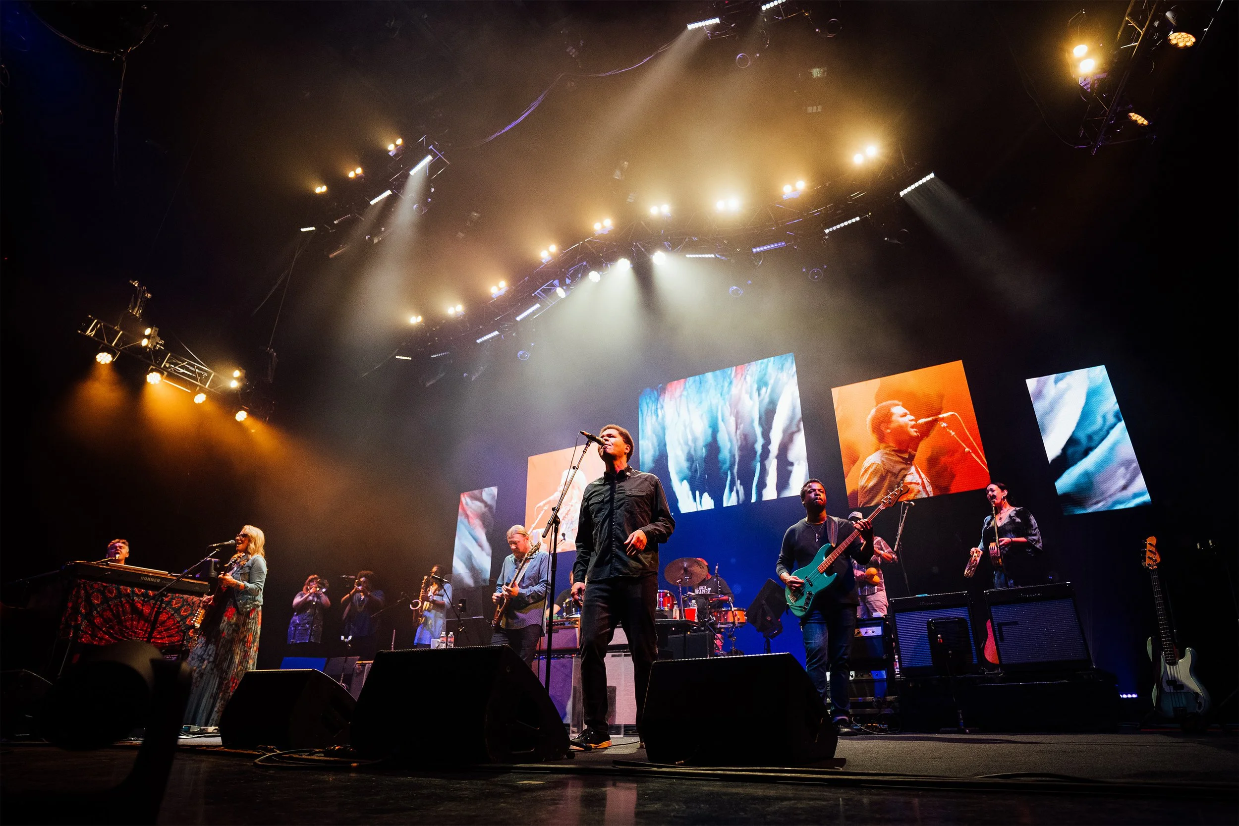 Band performing on stage with multiple musicians, colorful digital screens in the background, and stage lighting.