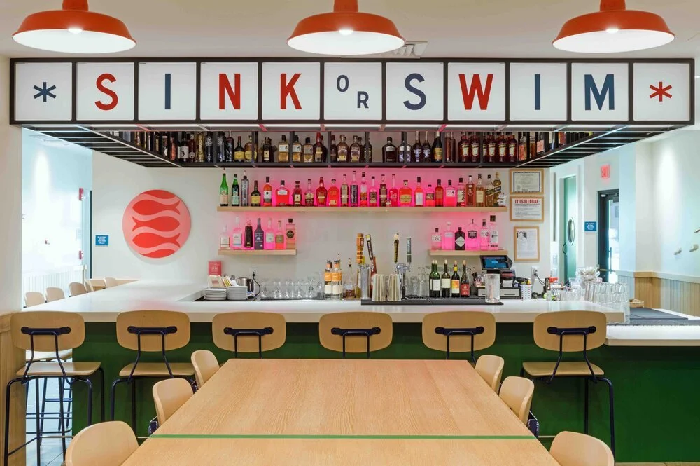 Interior of a modern bar with the words 'Sink or Swim' displayed above. The bar has stools, liquor bottles on shelves, and a countertop with glasses and bar tools.