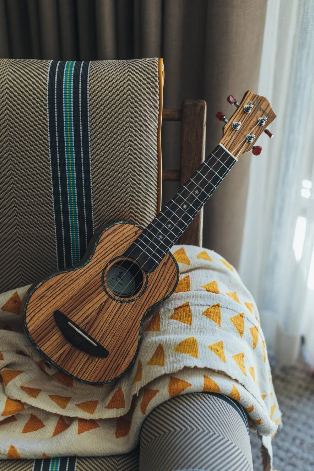 A small wooden ukulele resting on a patterned chair covered with a blanket featuring yellow triangles.