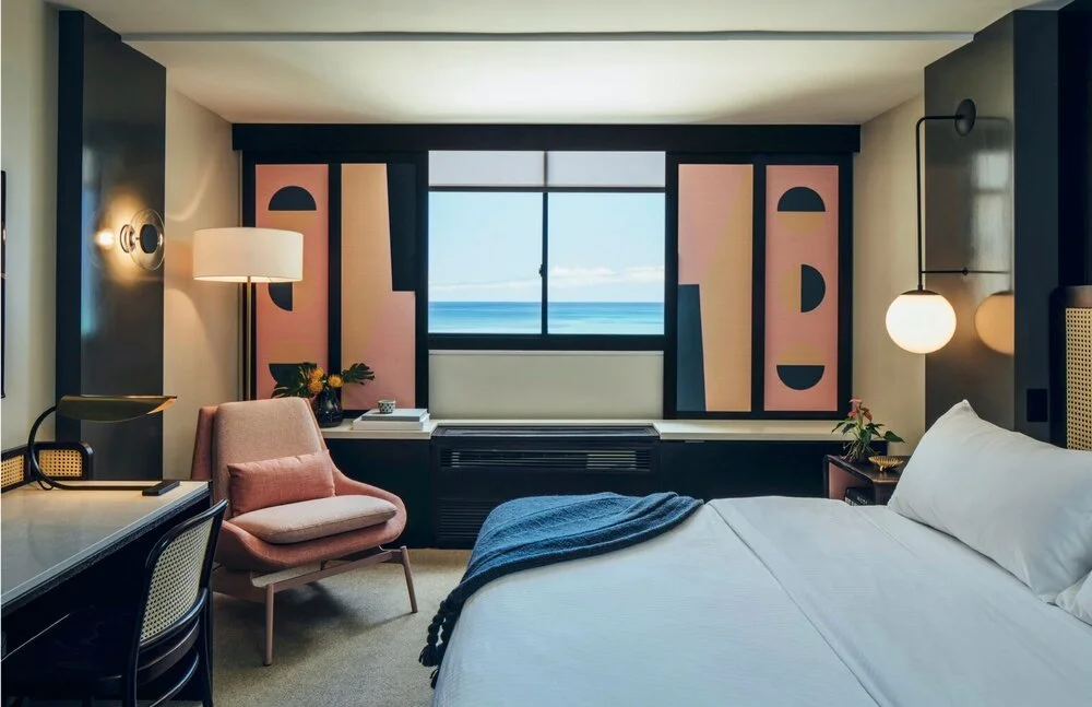 Modern hotel room with ocean view, featuring a bed, pink armchair, desk, and abstract artwork on window shades.